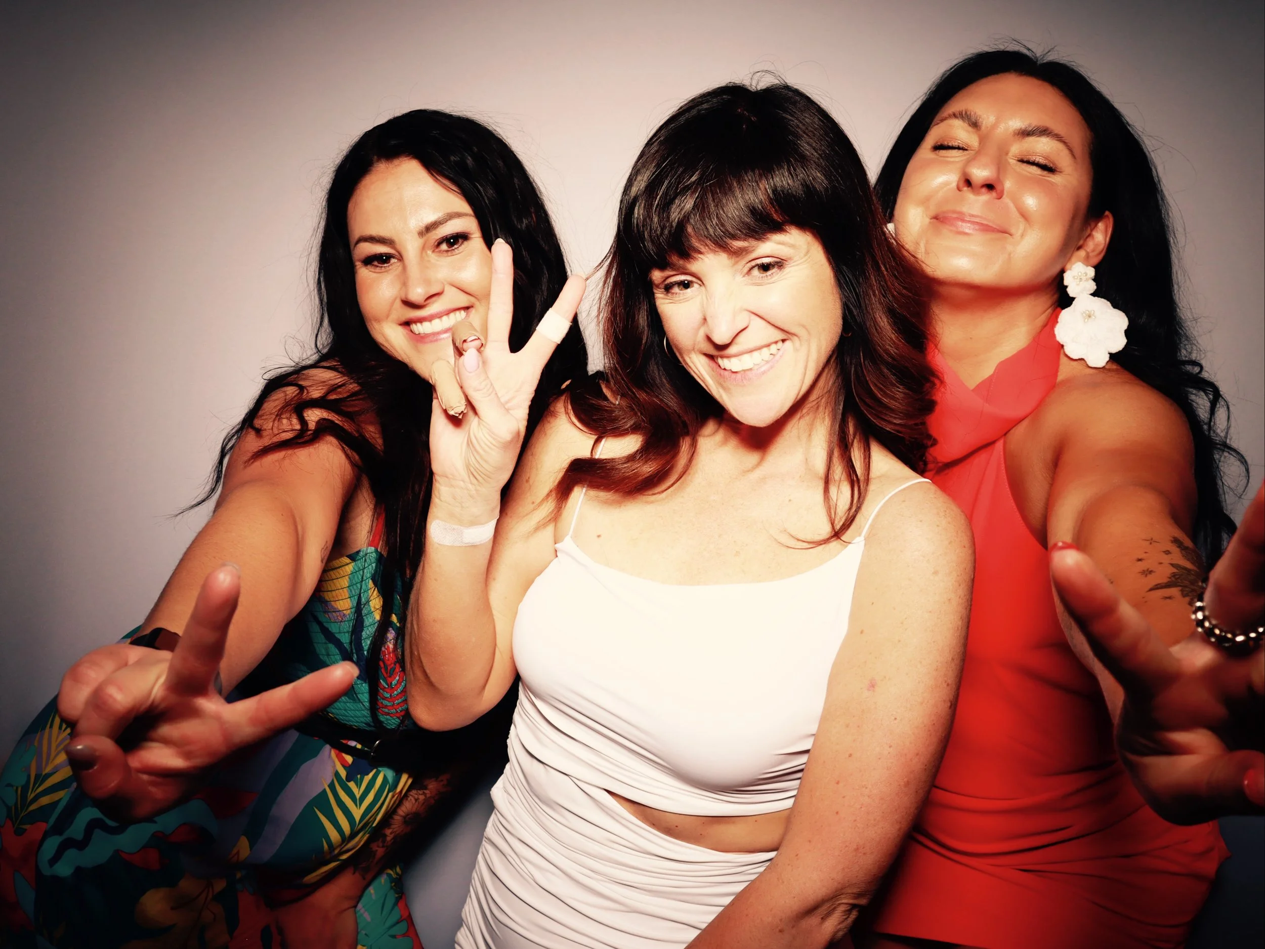 Three women taking a selfie together, smiling and making peace signs, against a neutral backdrop.