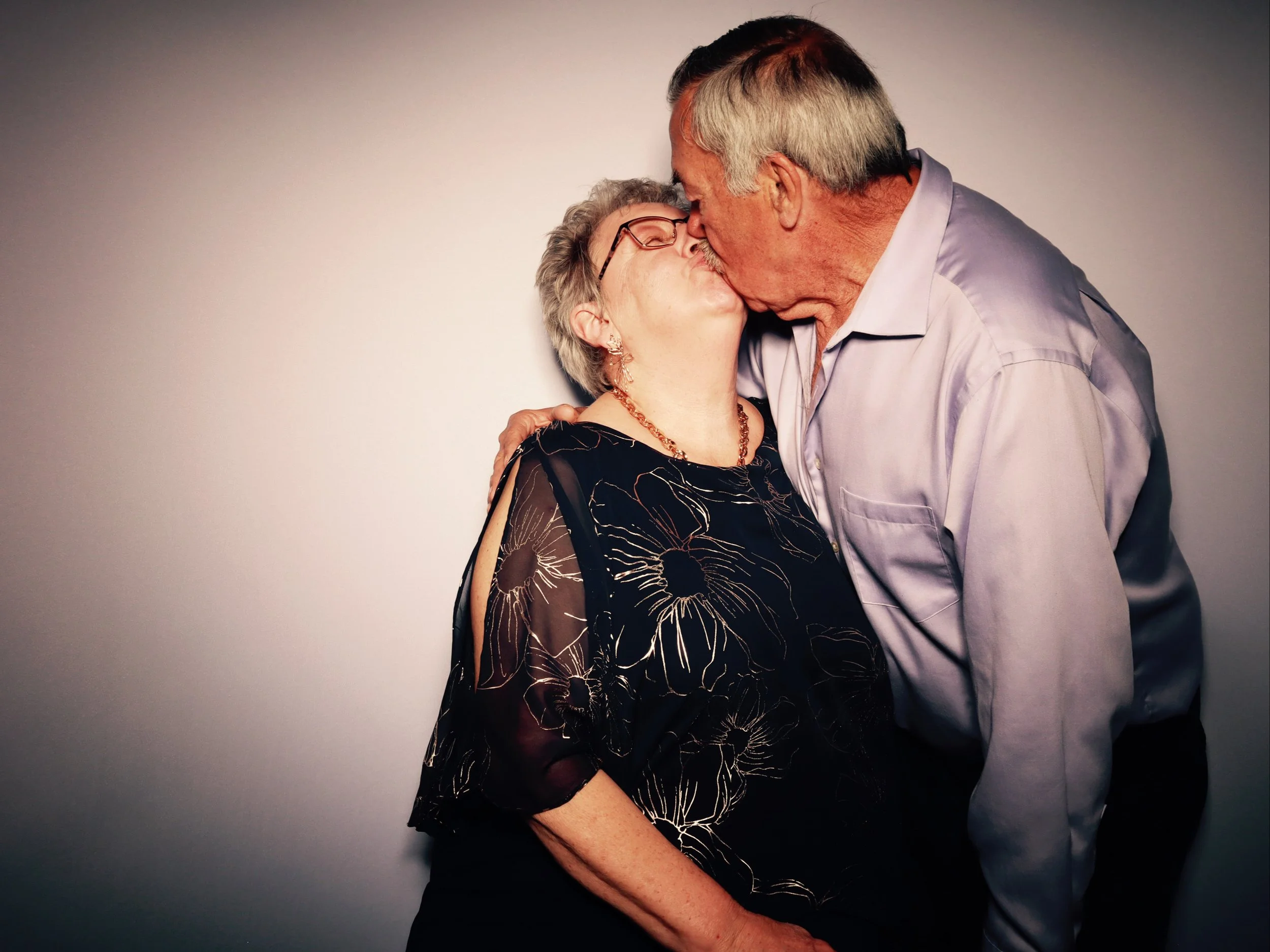 Older man and woman sharing a kiss, with the man holding the woman. They are dressed nicely and standing against a plain background.