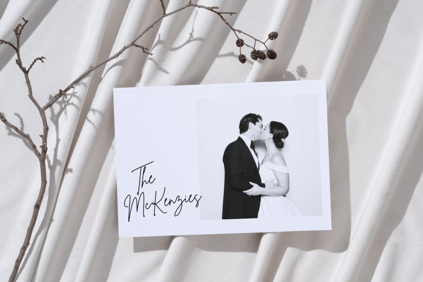 A black-and-white wedding photo of a bride and groom kissing, with the text 'The McKenzies' written in cursive on the photo, placed on a white fabric surface with a dried twig and small brown seed pods nearby.
