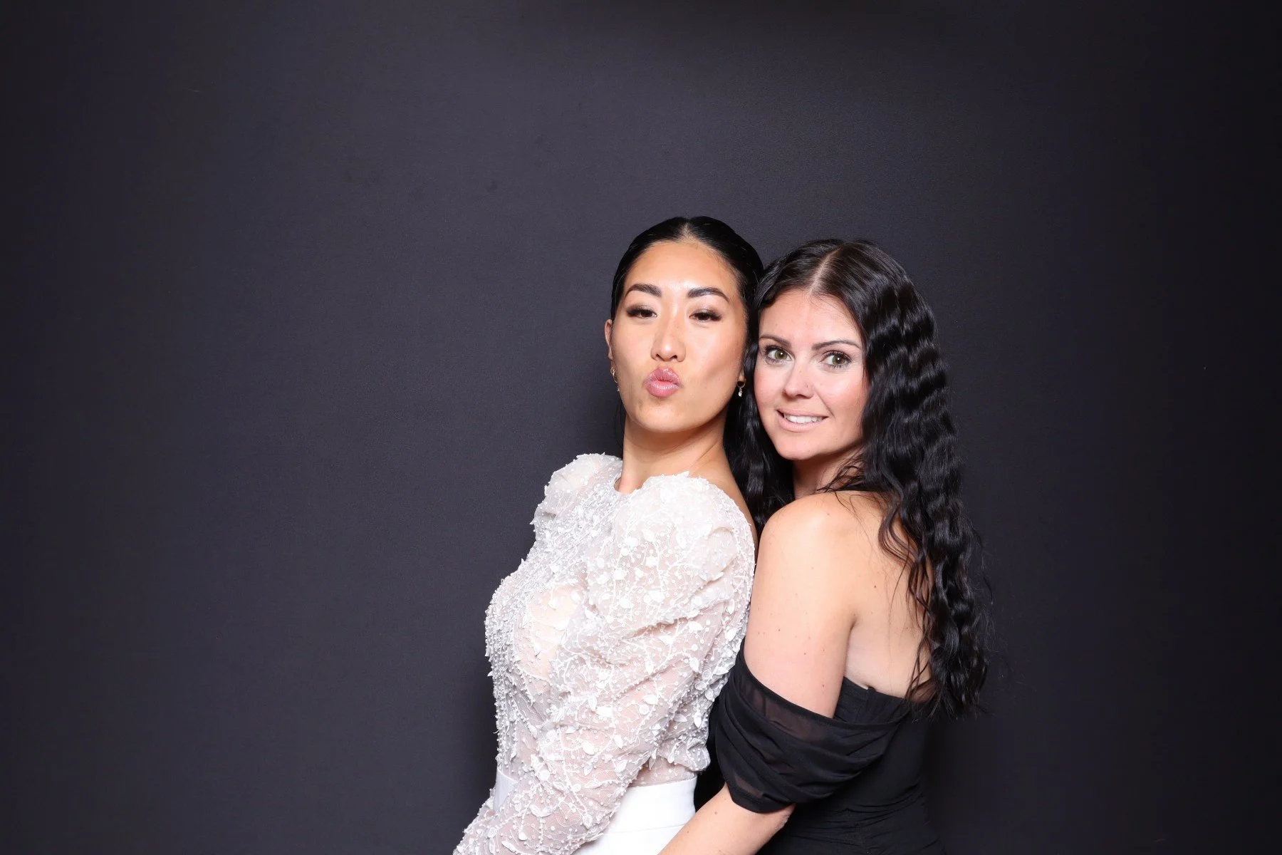 Two women with dark hair posing together against a black background, one in a white lace dress and the other in a black off-shoulder dress, smiling and making kissy faces.