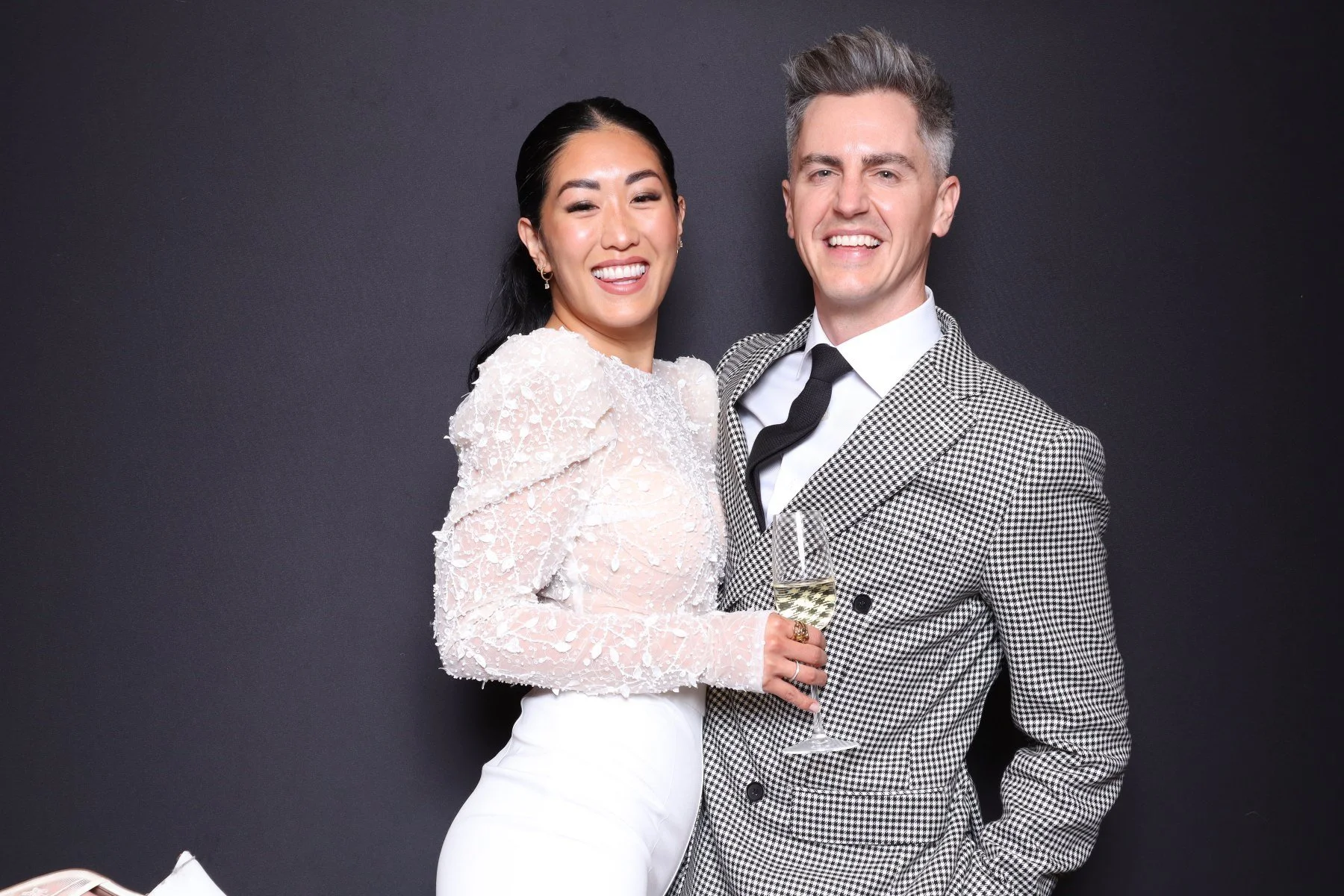 A happy couple dressed in formal attire, holding a glass of champagne, standing against a dark background.