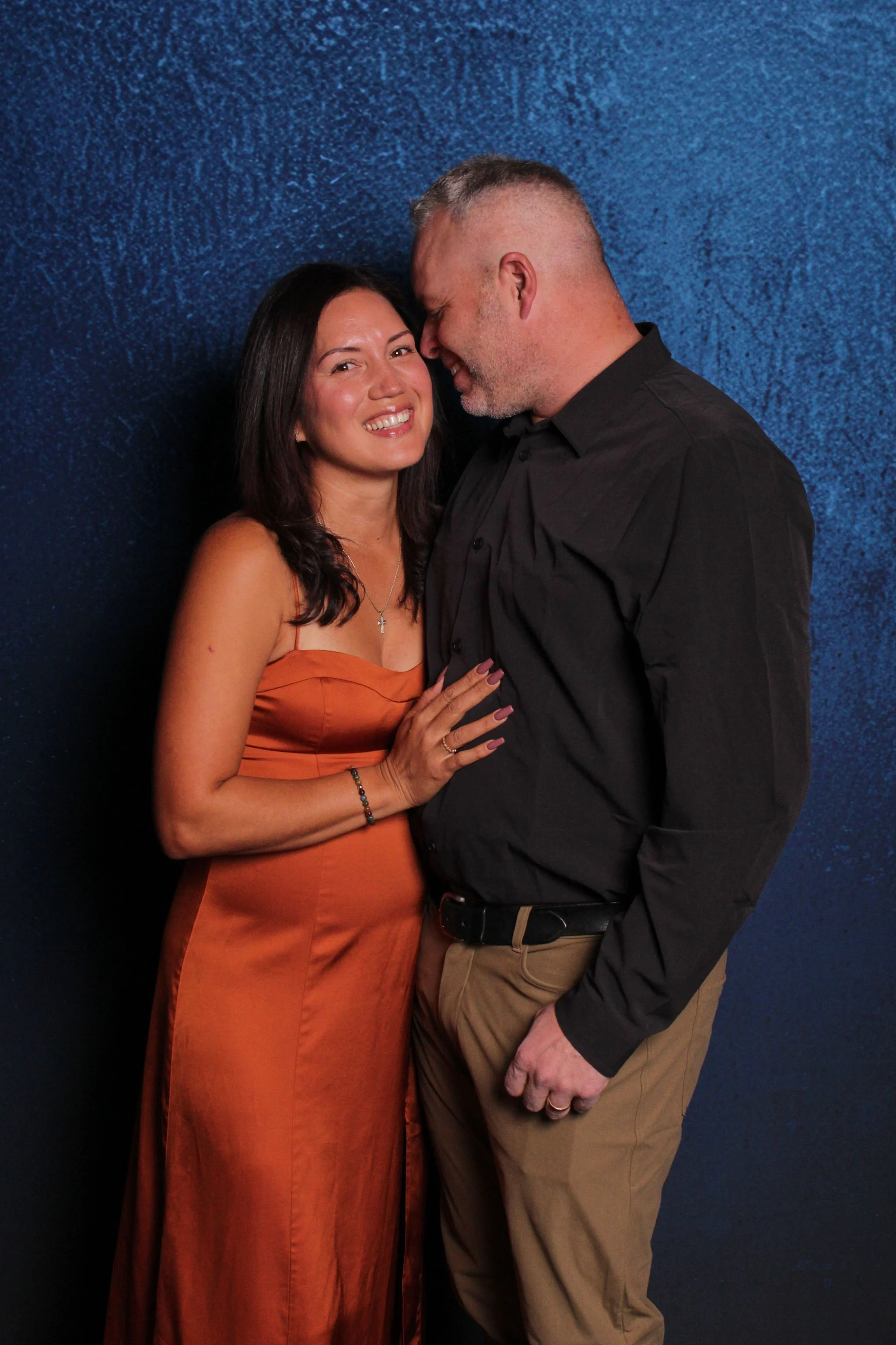 A smiling woman in a rust-colored dress and a man with a beard and gray hair in a black shirt and khaki pants share an intimate moment against a dark blue textured background.