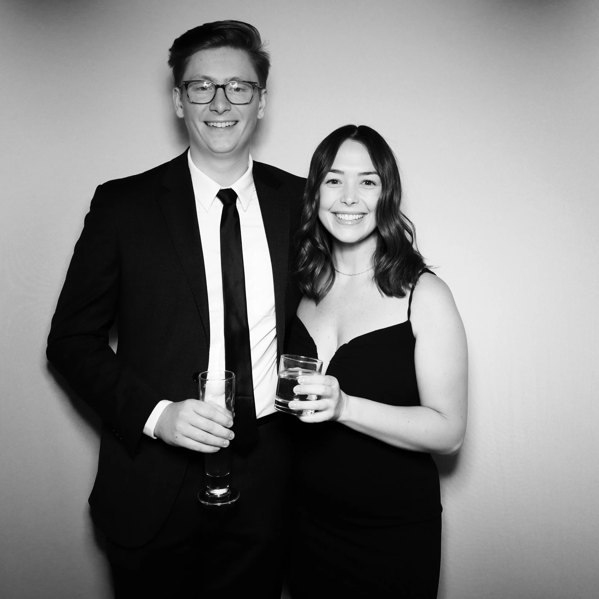A young man in a suit and a young woman in a dress smiling and holding drinks, standing together against a plain background.
