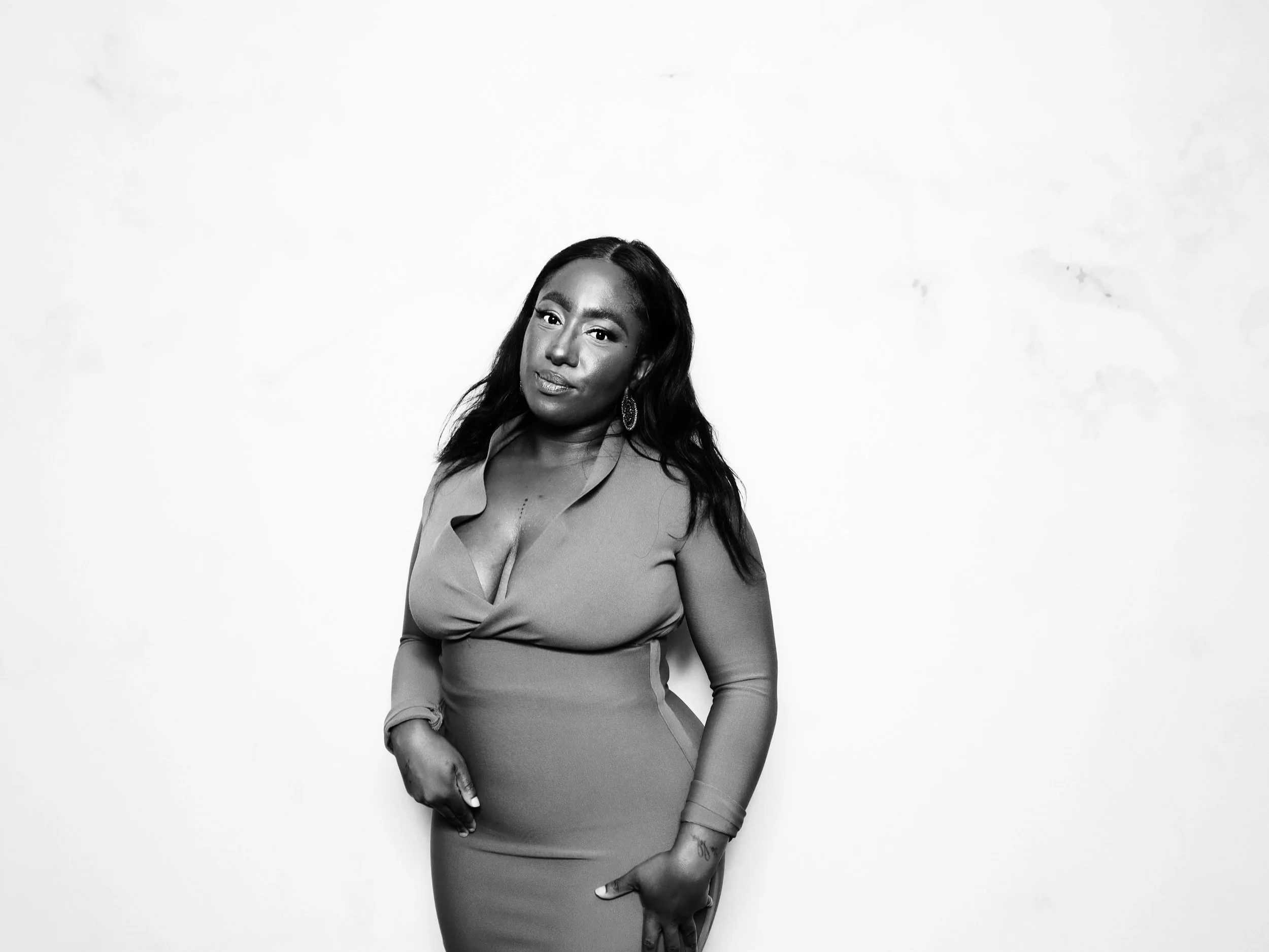 Black and white portrait of a woman with long hair, wearing a fitted dress and earrings, standing against a plain white background.