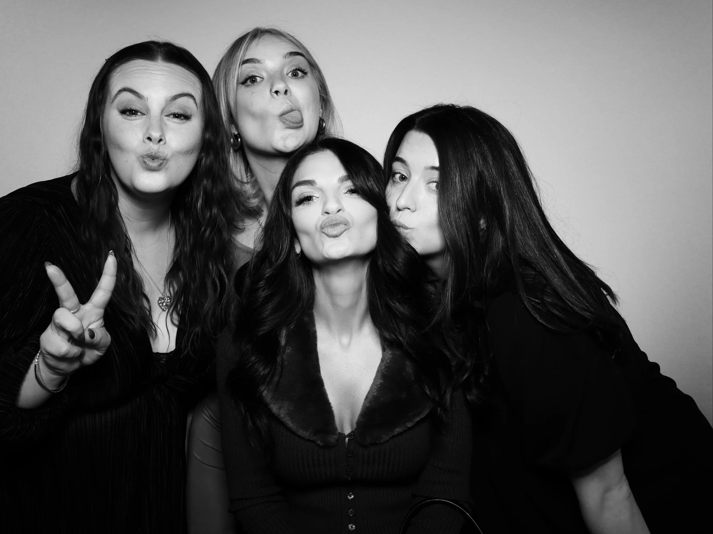 Four women posing for a photo in black and white, making playful facial expressions and gestures, against a plain background.