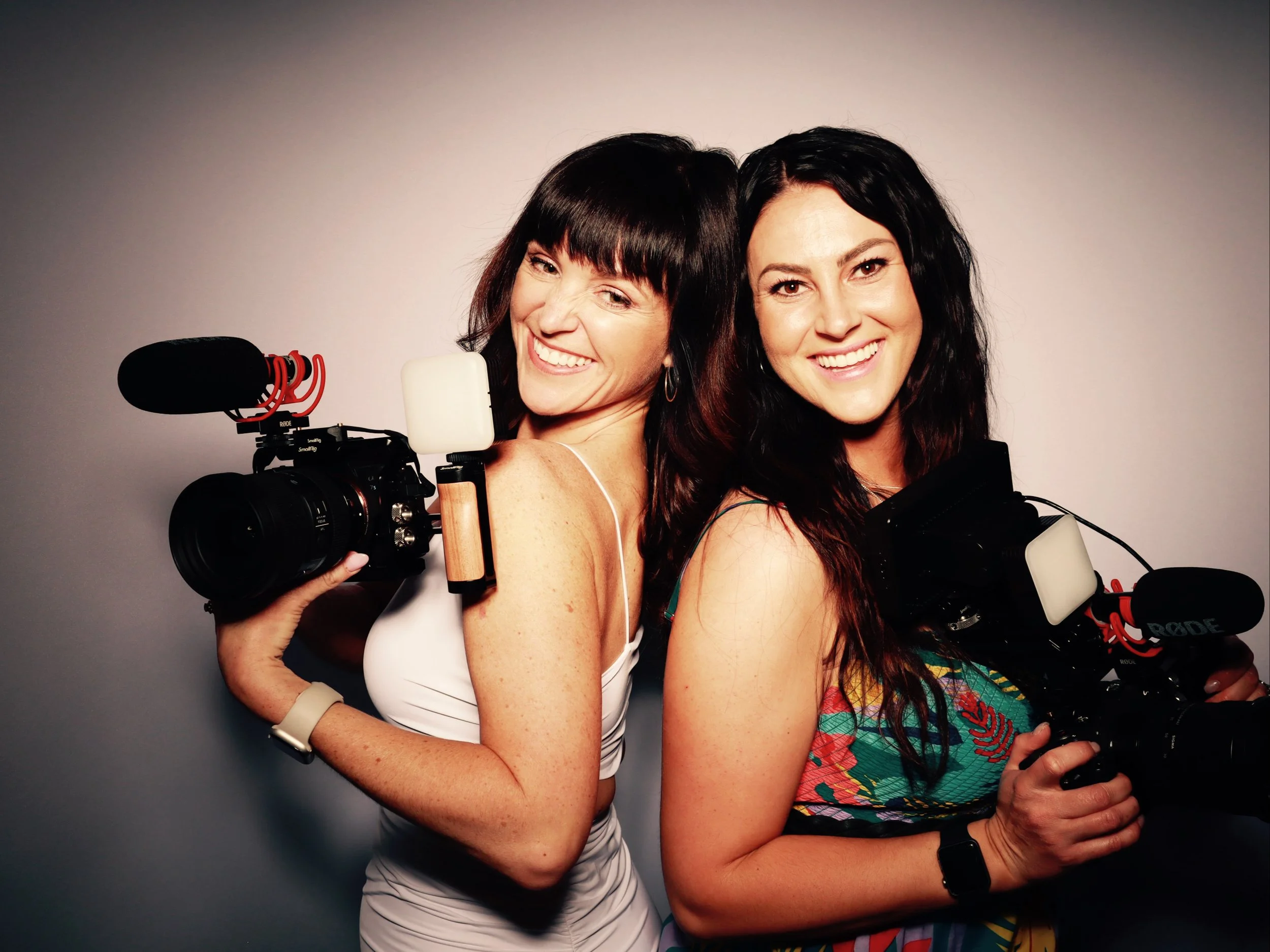 Two women smiling and holding video cameras with microphones, standing back to back against a plain background.