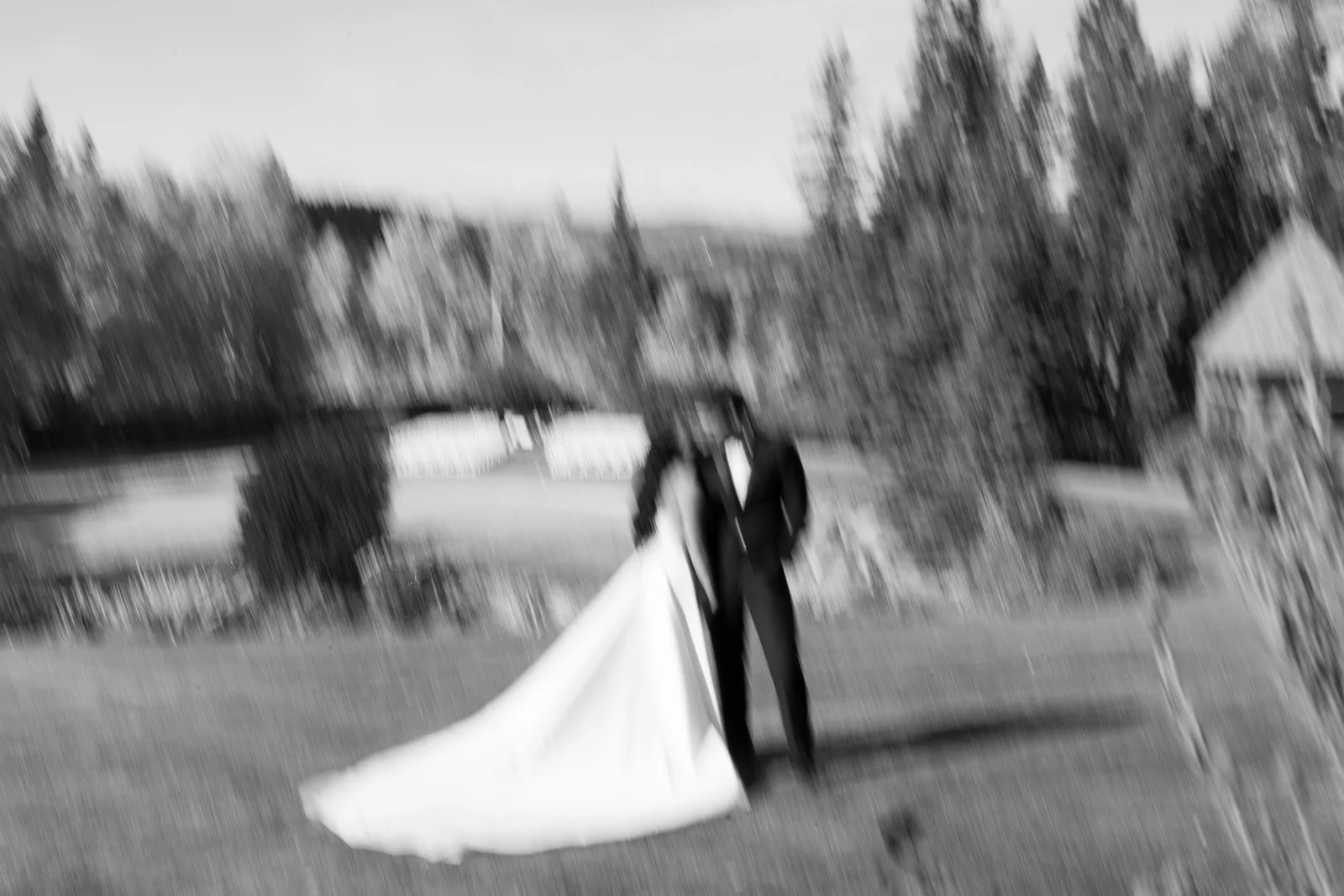A blurry black and white photo of a bride and groom standing outside near a tree and a house, dressed in wedding attire.