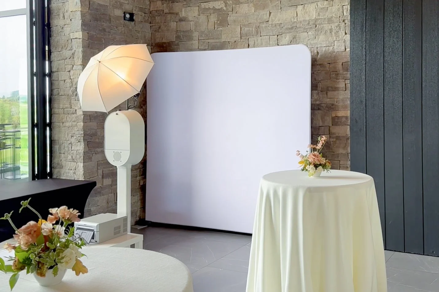 Interior of a modern room with a textured brick wall, large windows, a round table with a floral centerpiece, and a photo booth with a white backdrop, umbrella, and lighting equipment used for taking pictures.
