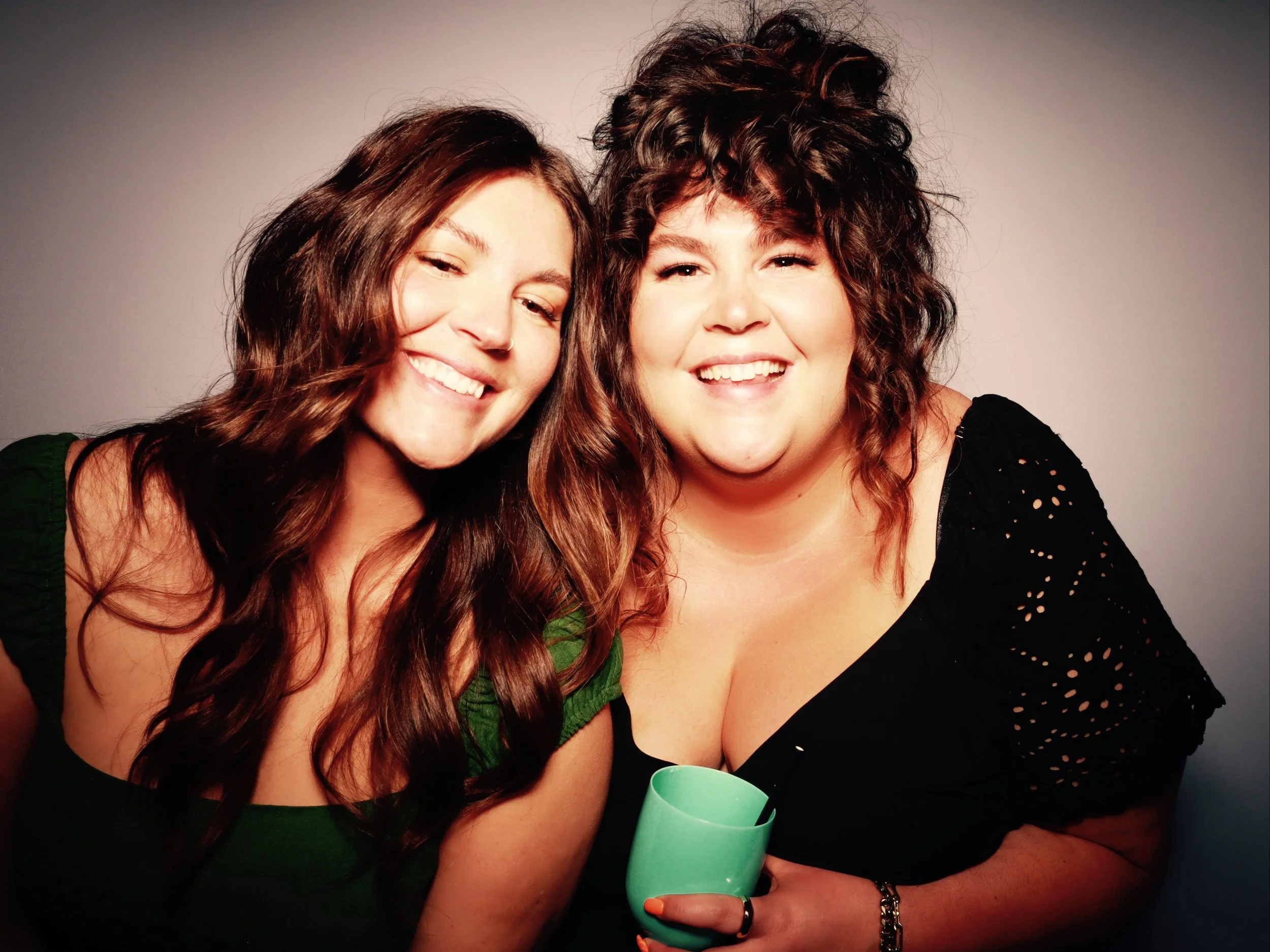Two women smiling, one holding a green cup, posing closely against a neutral background.