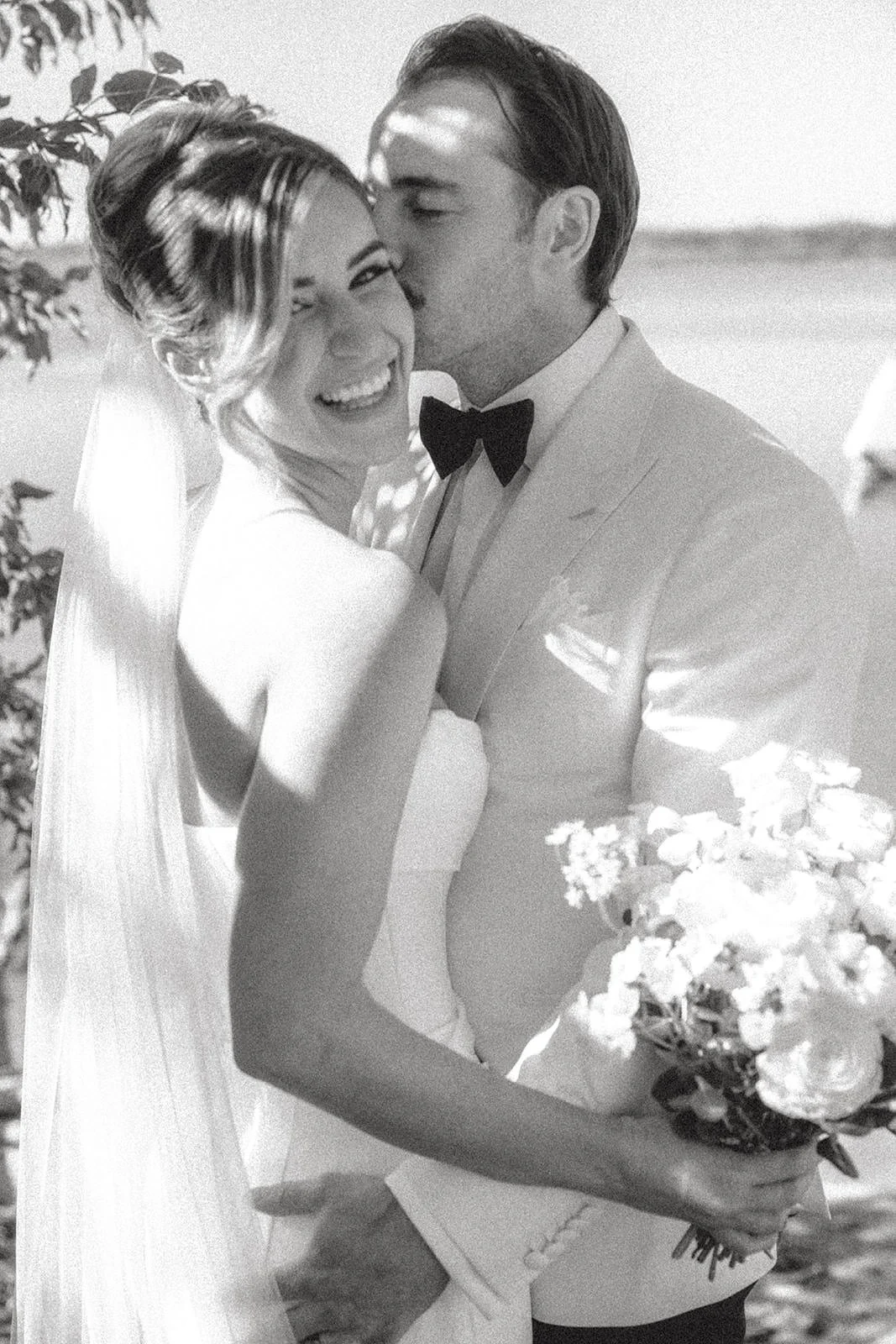 A black-and-white photo of a bride and groom on their wedding day, sharing a joyful moment, with the groom kissing the bride's temple and the bride smiling.