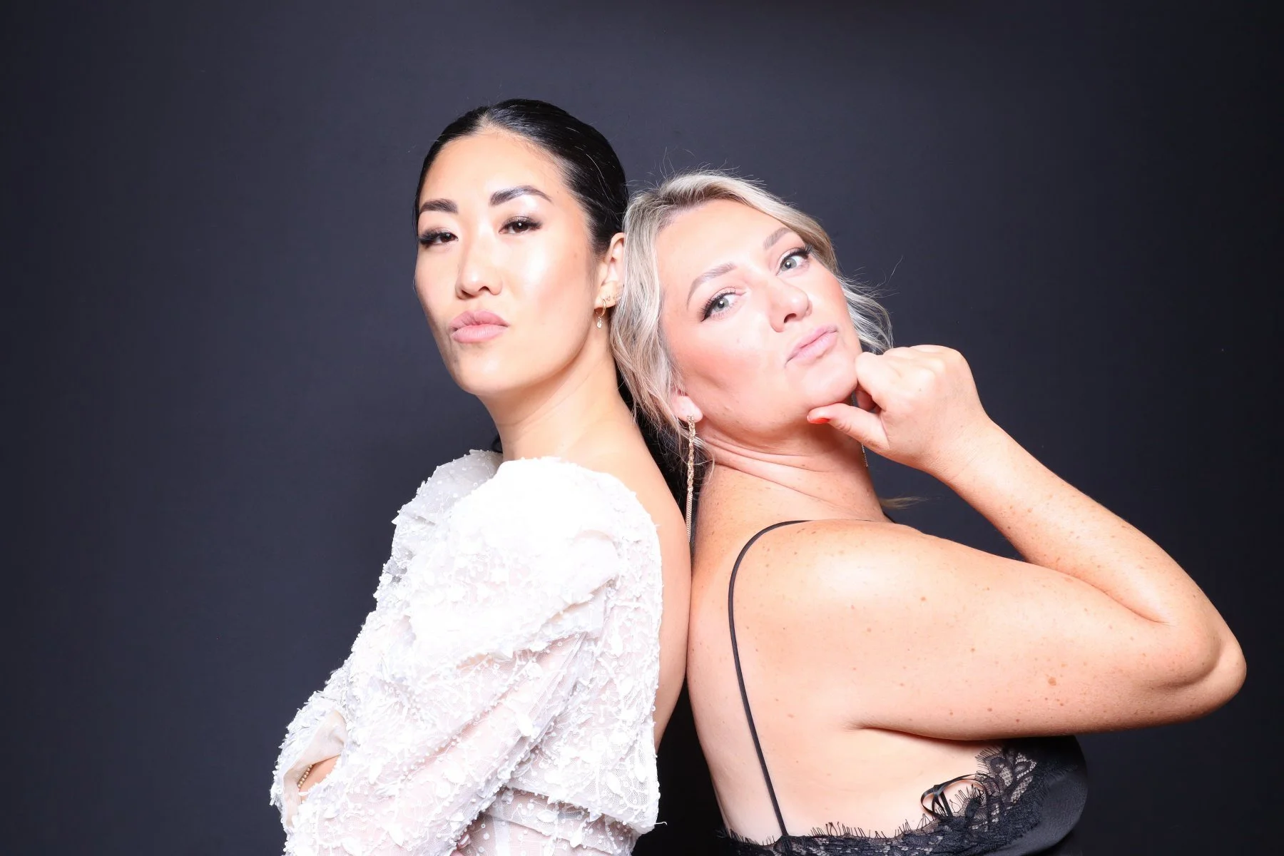 Two women standing back to back with serious expressions, one with dark hair and the other with blonde hair, against a dark background.