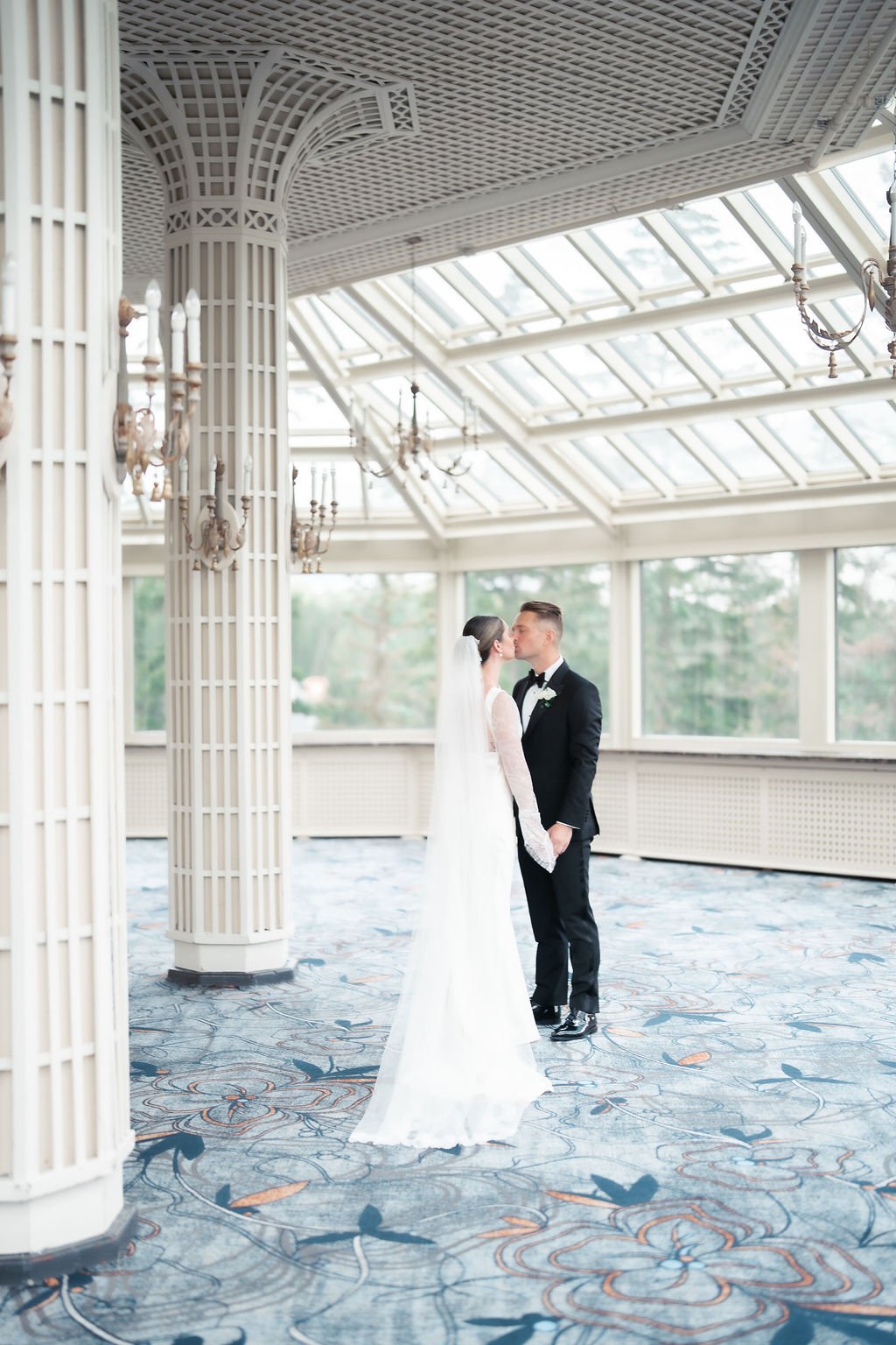 Bride and groom kissing in a bright indoor setting with large glass ceiling and ornate chandeliers.