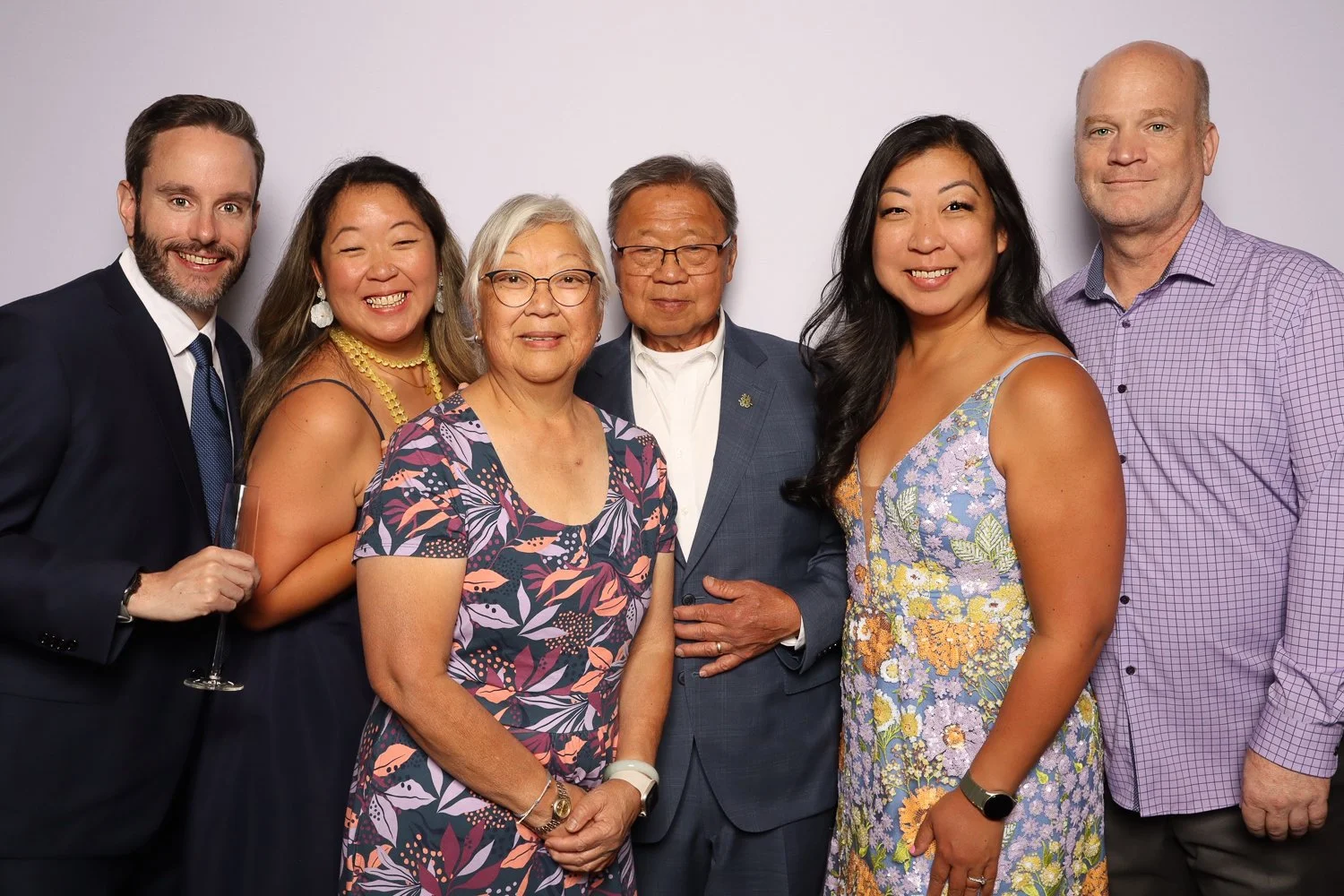 Group of seven diverse adults, three men and four women, dressed in formal and semi-formal attire, standing together and smiling against a plain background.