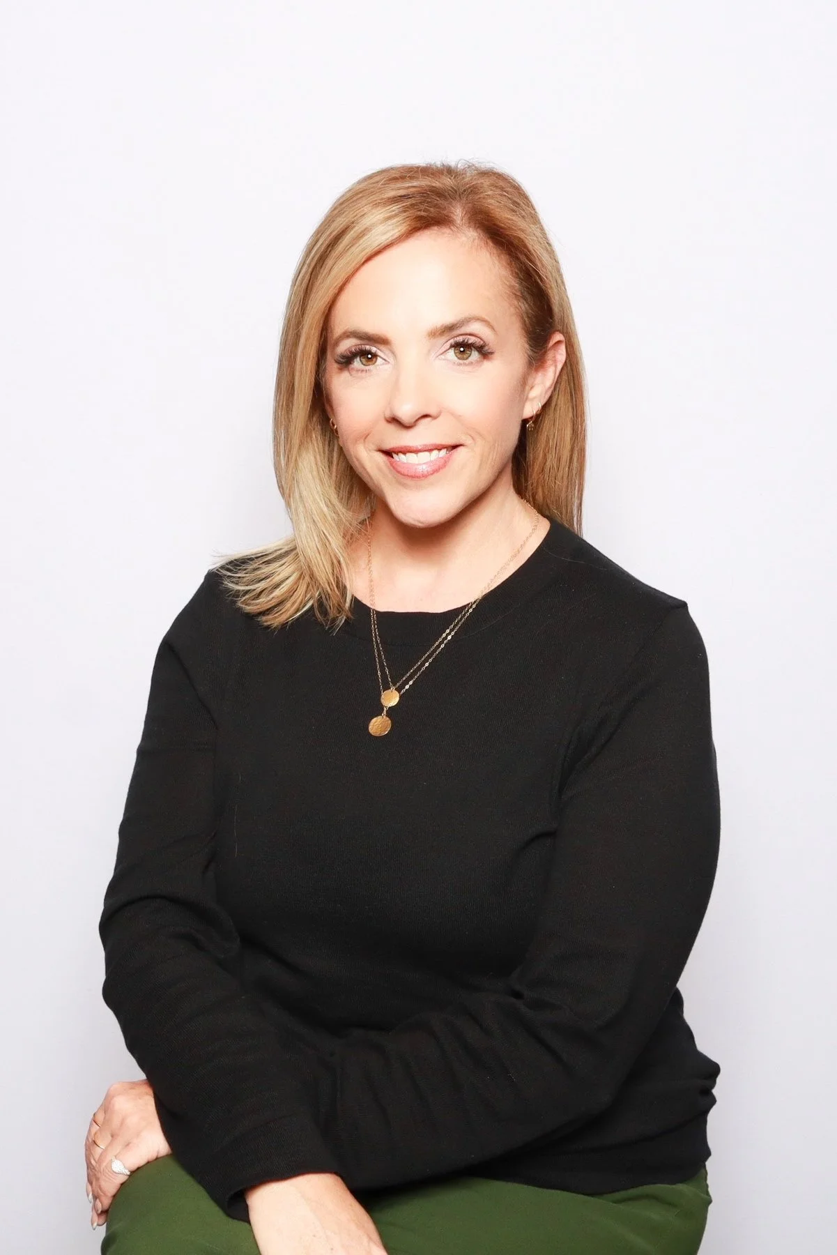 A woman with blonde hair smiling, wearing a black top and gold necklace, sitting against a plain white background.