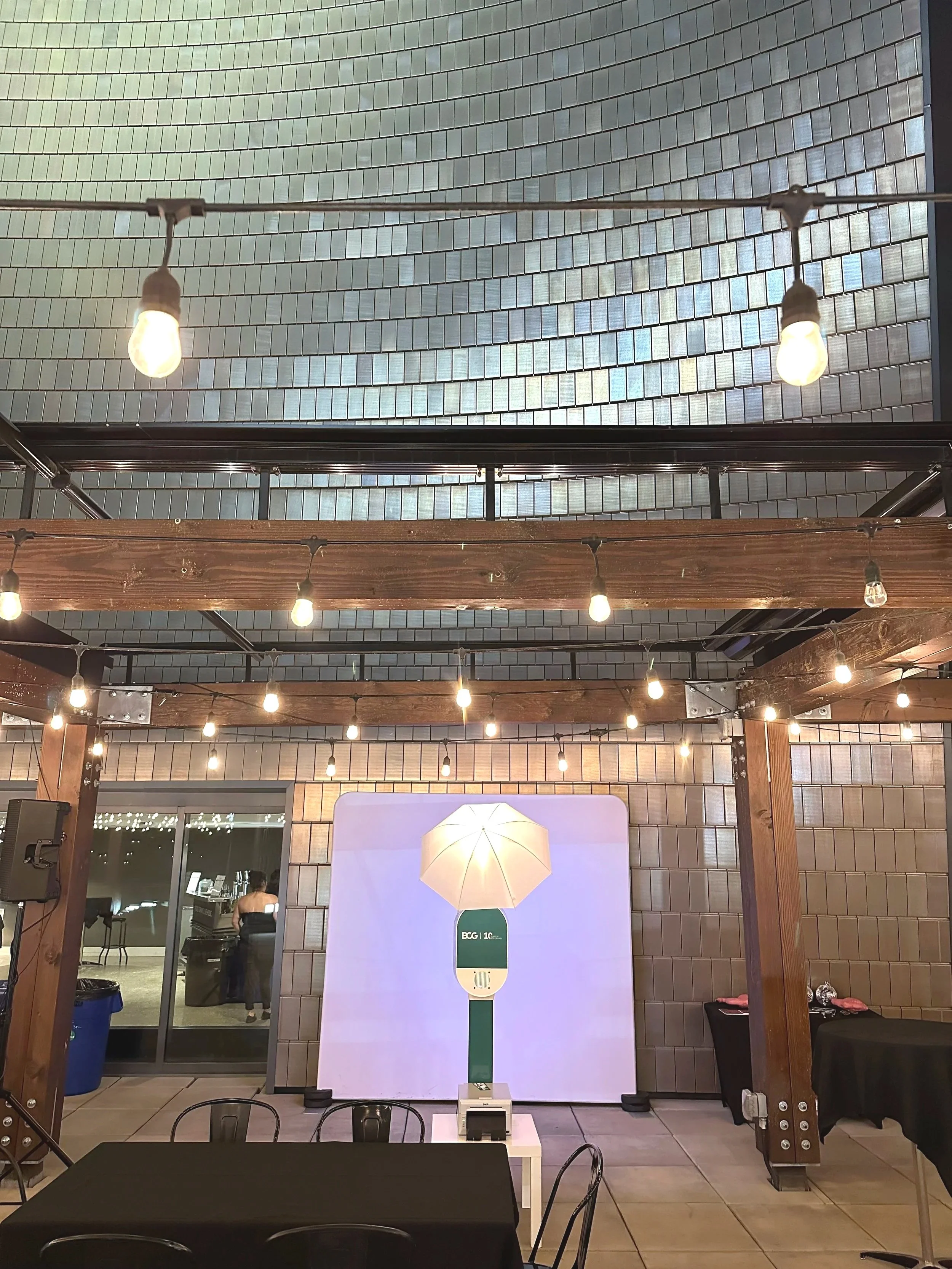 Indoor event space with string lights, wooden beams, a large illuminated umbrella prop, and a backdrop with a green logo, brick and tiled walls, and a person in the background near a glass door.