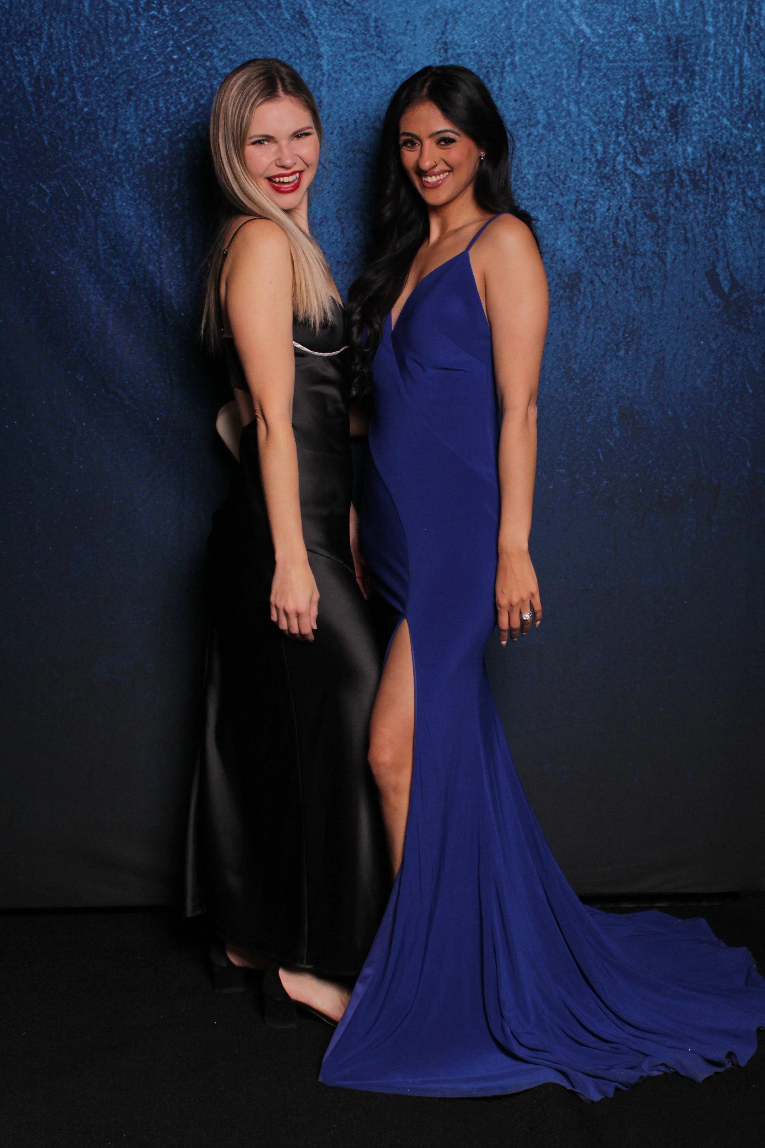 Two women dressed in elegant gowns posing together against a dark blue textured background, smiling at the camera.