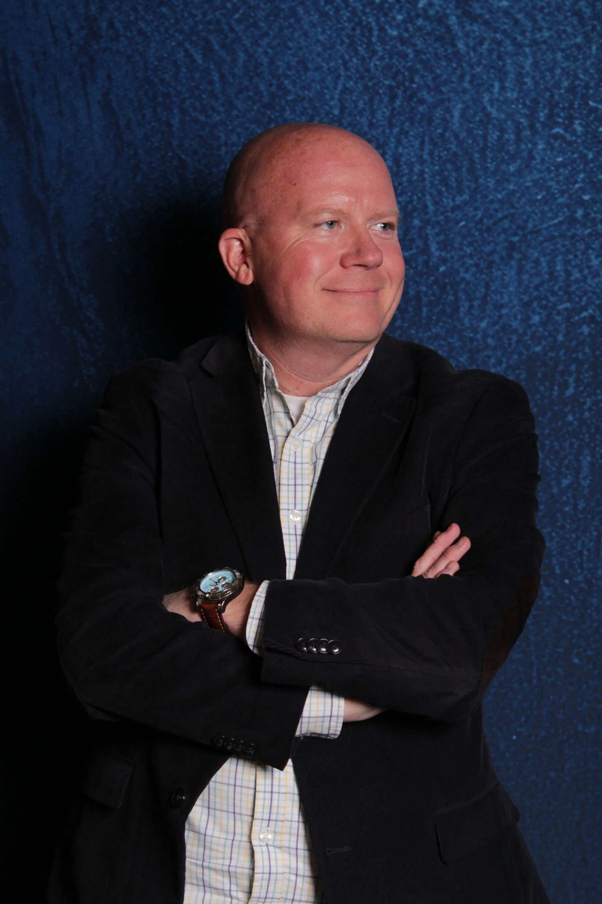 A man with a bald head wearing a black blazer and a checkered shirt, crossing his arms, standing against a textured blue background.