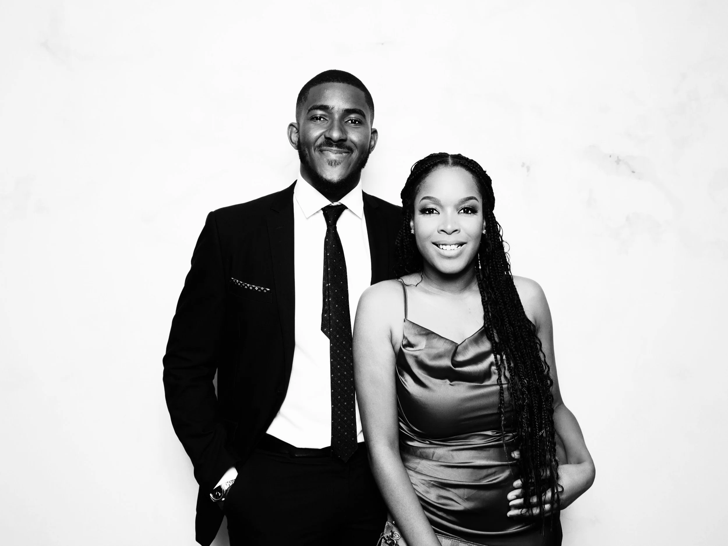 A black and white photo of a smiling man and woman standing close together against a plain background. The man is wearing a suit and tie, and the woman is wearing a satin dress with spaghetti straps.