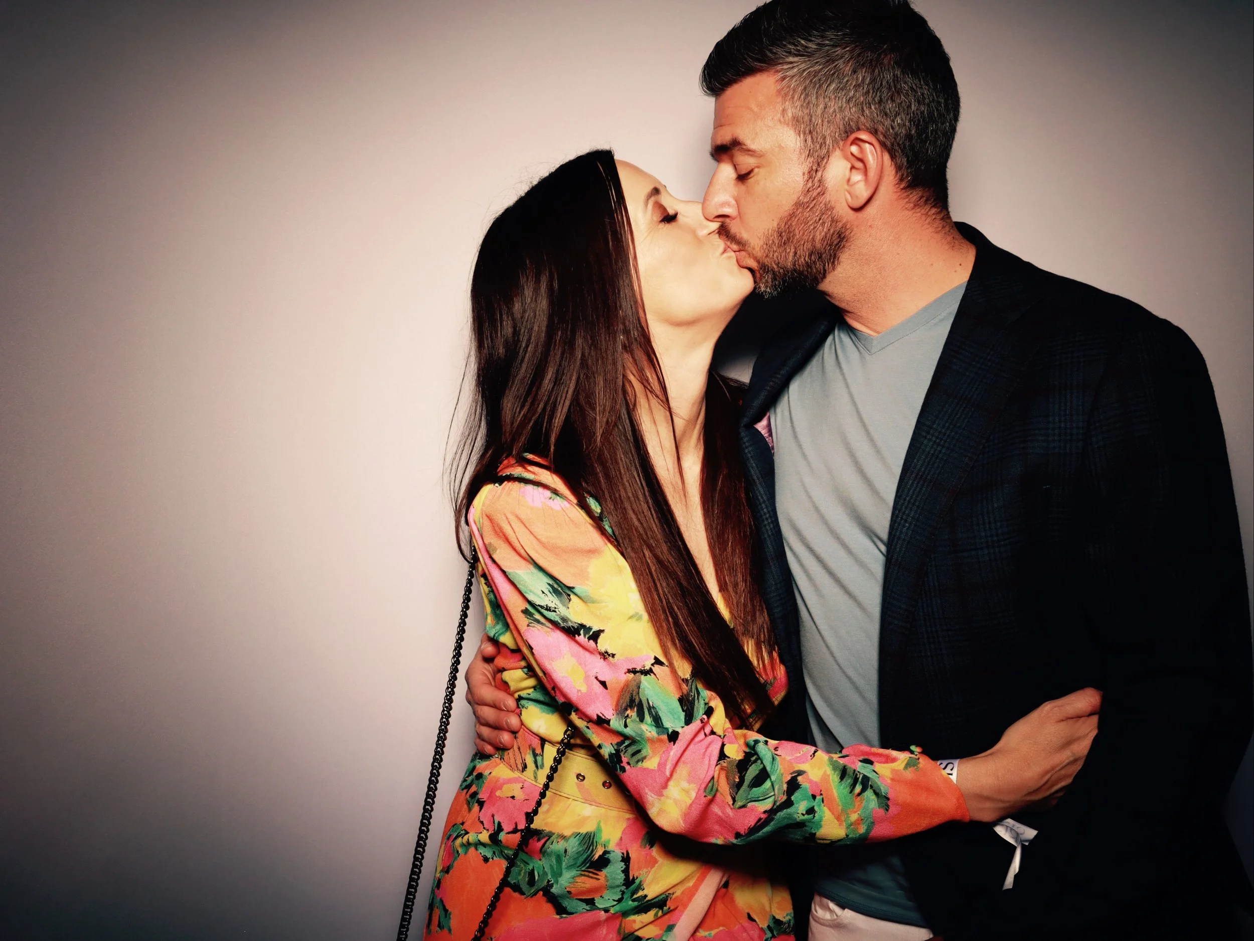 A man and woman are sharing a kiss, with their eyes closed, against a plain background. The woman has long dark hair and is wearing a colorful floral dress, while the man has short dark hair, a beard, and is dressed in a gray t-shirt and dark blazer.