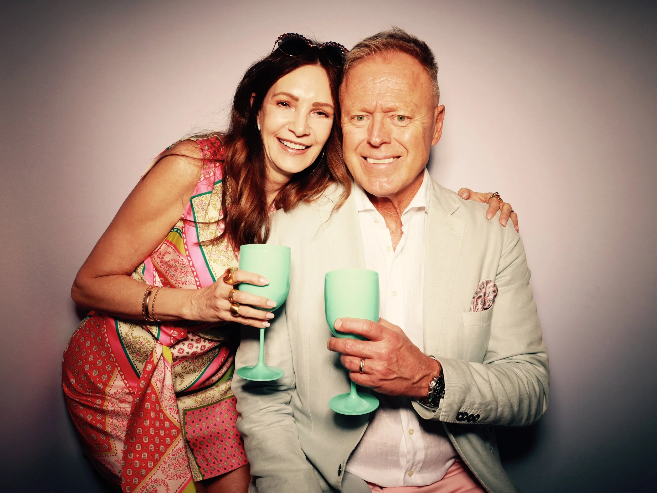 A smiling woman and man holding pastel green cocktail glasses, standing close together against a plain gray background.
