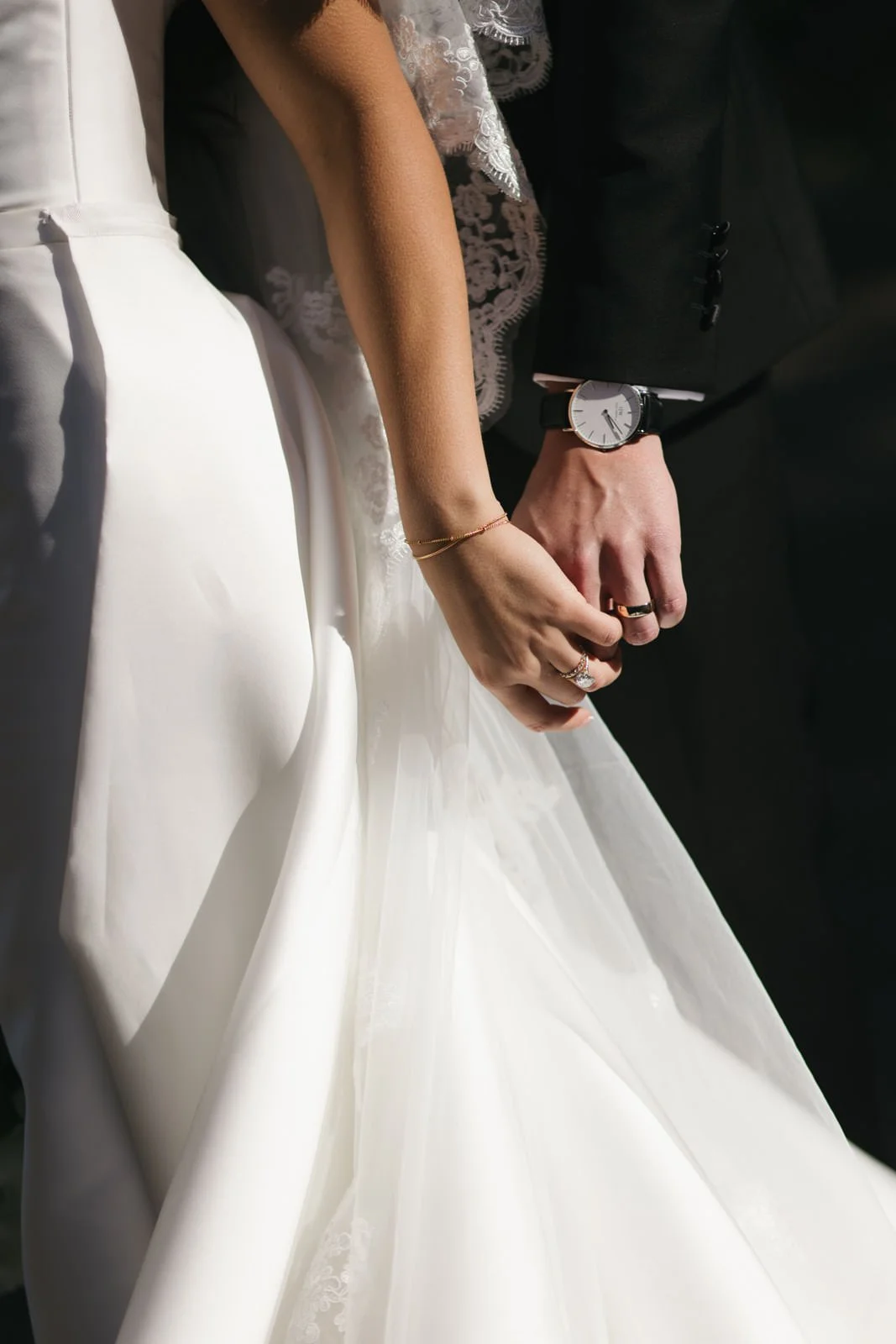 Close-up of a bride and groom holding hands during a wedding ceremony, focusing on their wedding rings and the bride's wedding dress.