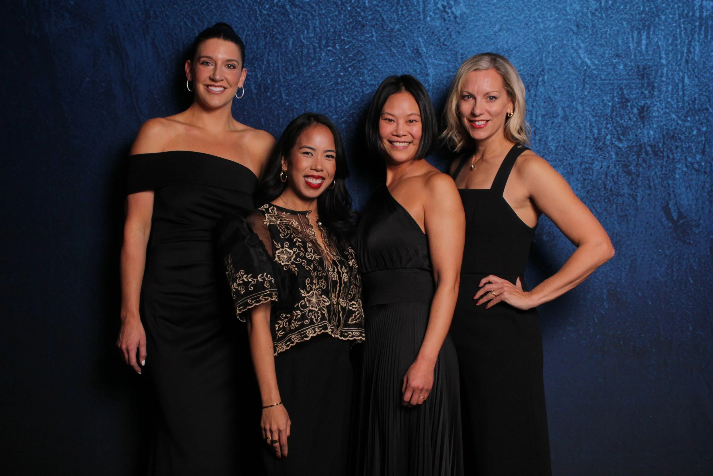 Four women dressed in black formal attire standing against a dark blue textured backdrop, smiling at the camera.