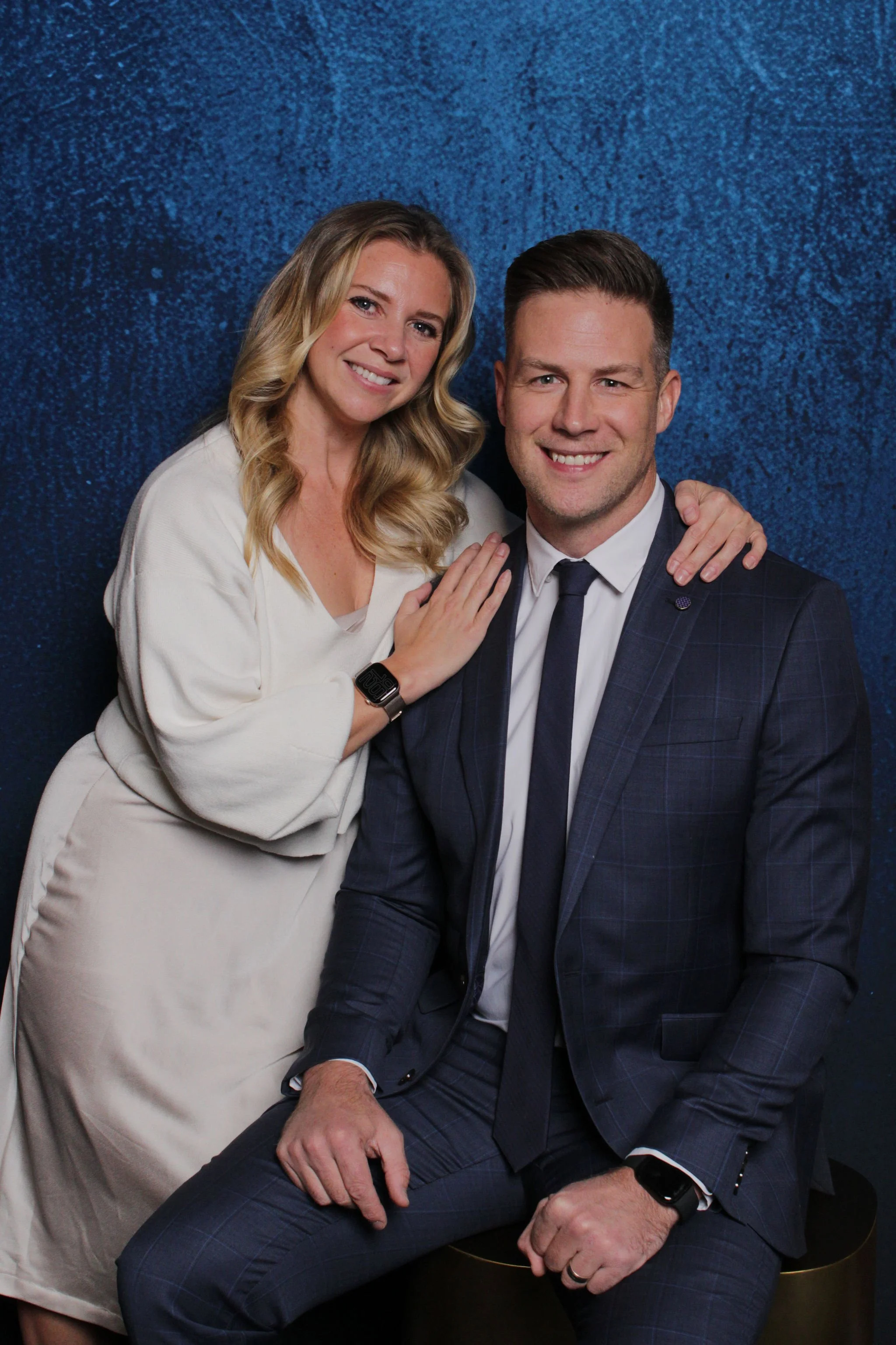 A woman with blonde hair and a man with short brown hair smiling at the camera. The woman has her hand on the man's shoulder, and the man is sitting, wearing a dark blue suit, white shirt, and dark tie. They are posing against a blue textured backgro