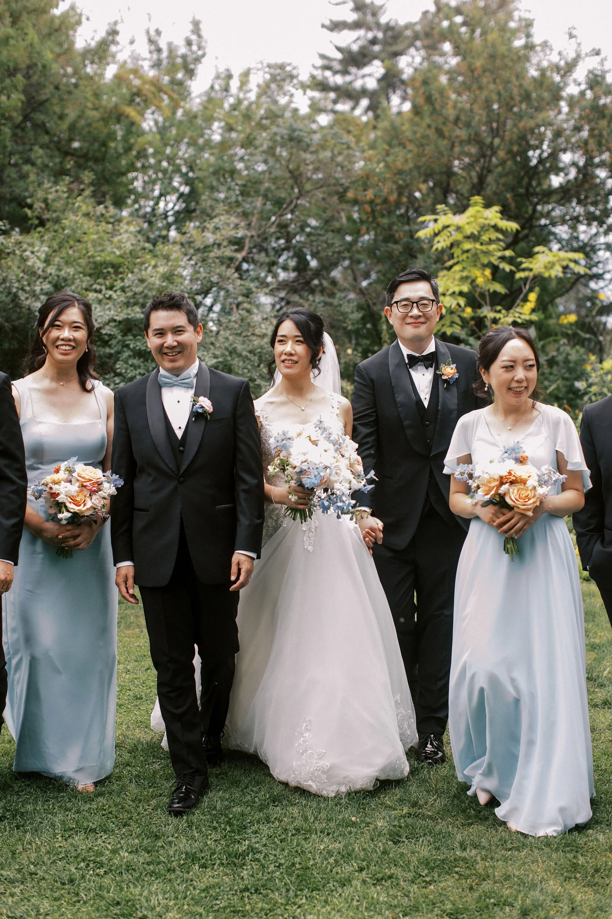 Group of six wedding guests, including a bride in a white wedding dress holding a bouquet, standing outdoors on grass with trees in the background.