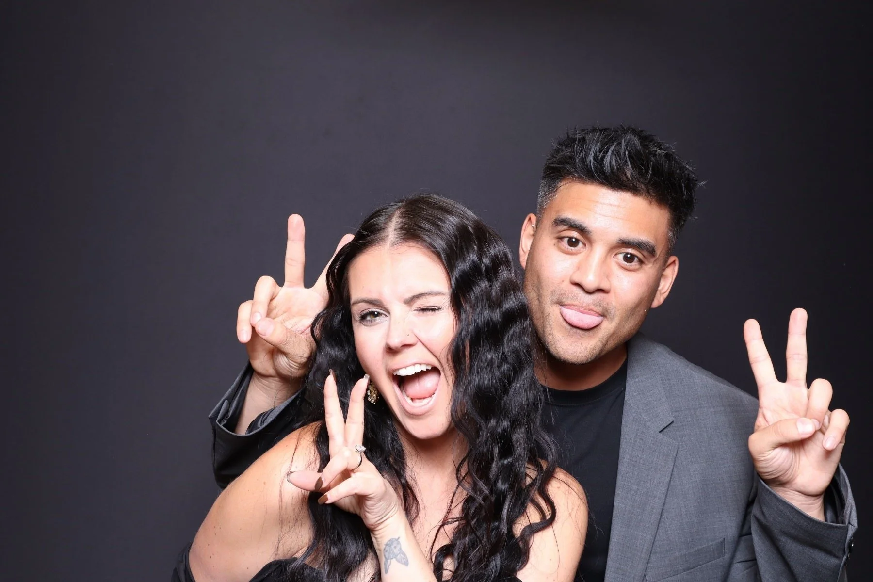Two people posing playfully against a black background, making peace signs and sticking out their tongues.