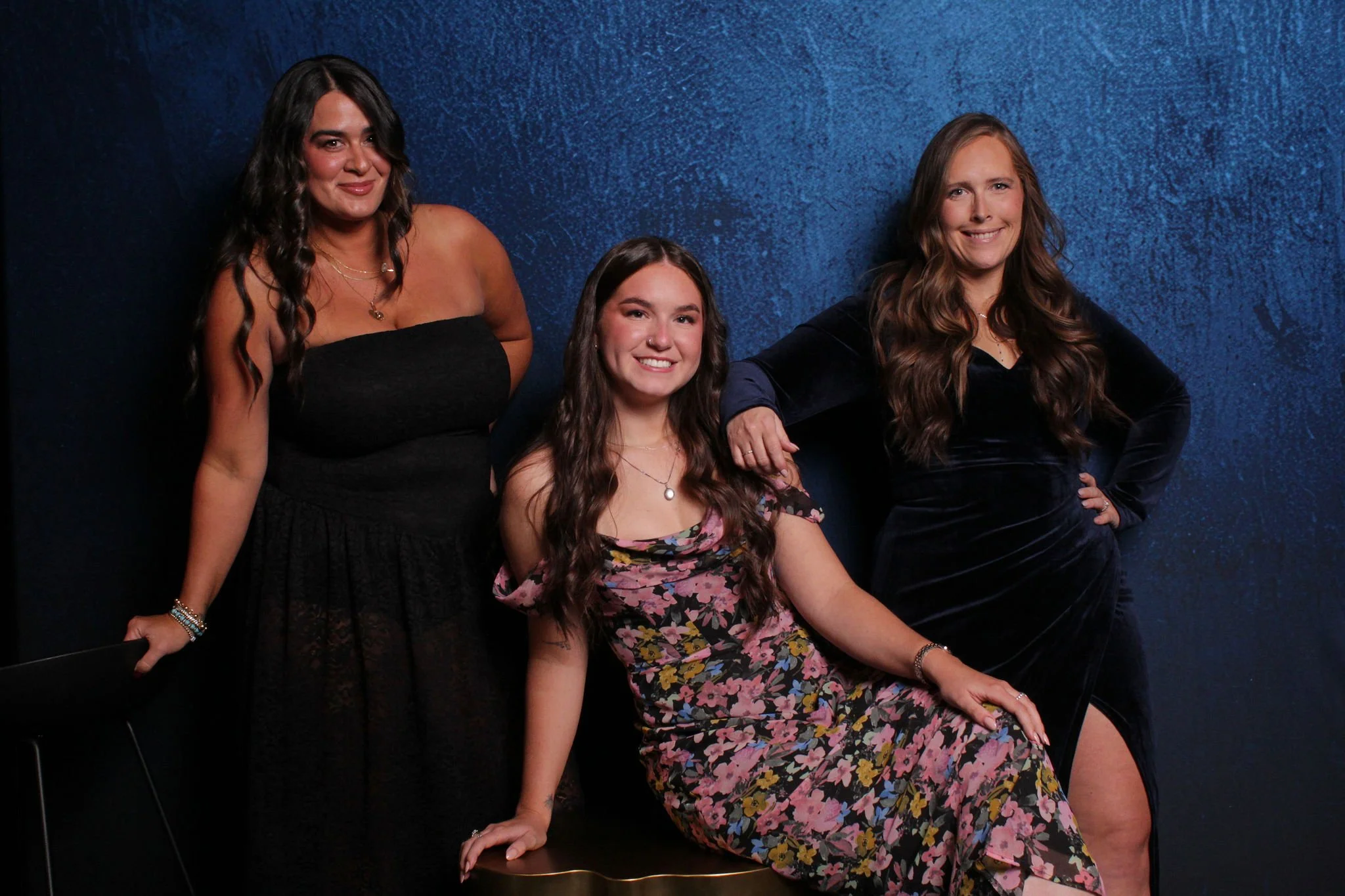 Three women posing against a dark blue textured background, with one seated on a gold cushion and two standing beside her. The woman on the left has long dark hair and is wearing a strapless black dress. The woman in the middle has long wavy brown ha