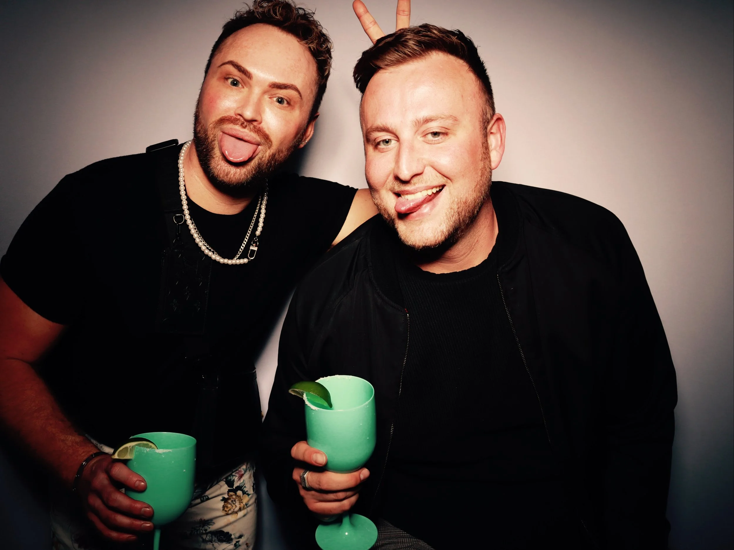 Two men celebrating with green cocktails, one making bunny ears behind the other, both sticking out their tongues and smiling.