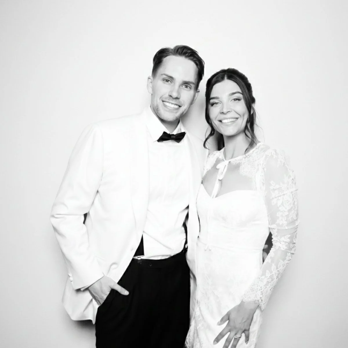 Black-and-white photo of a bride and groom smiling, dressed in wedding attire, standing close together against a plain background.