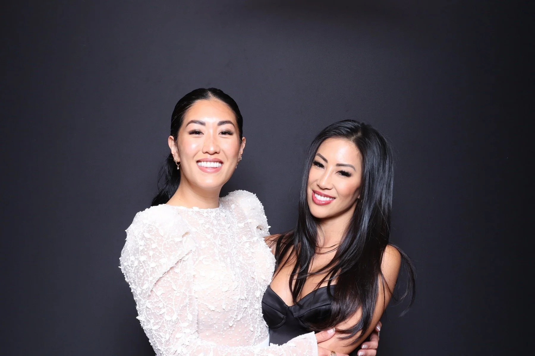 Two smiling women with dark hair and makeup, one wearing a white lace dress and the other wearing a black top, standing against a dark background.