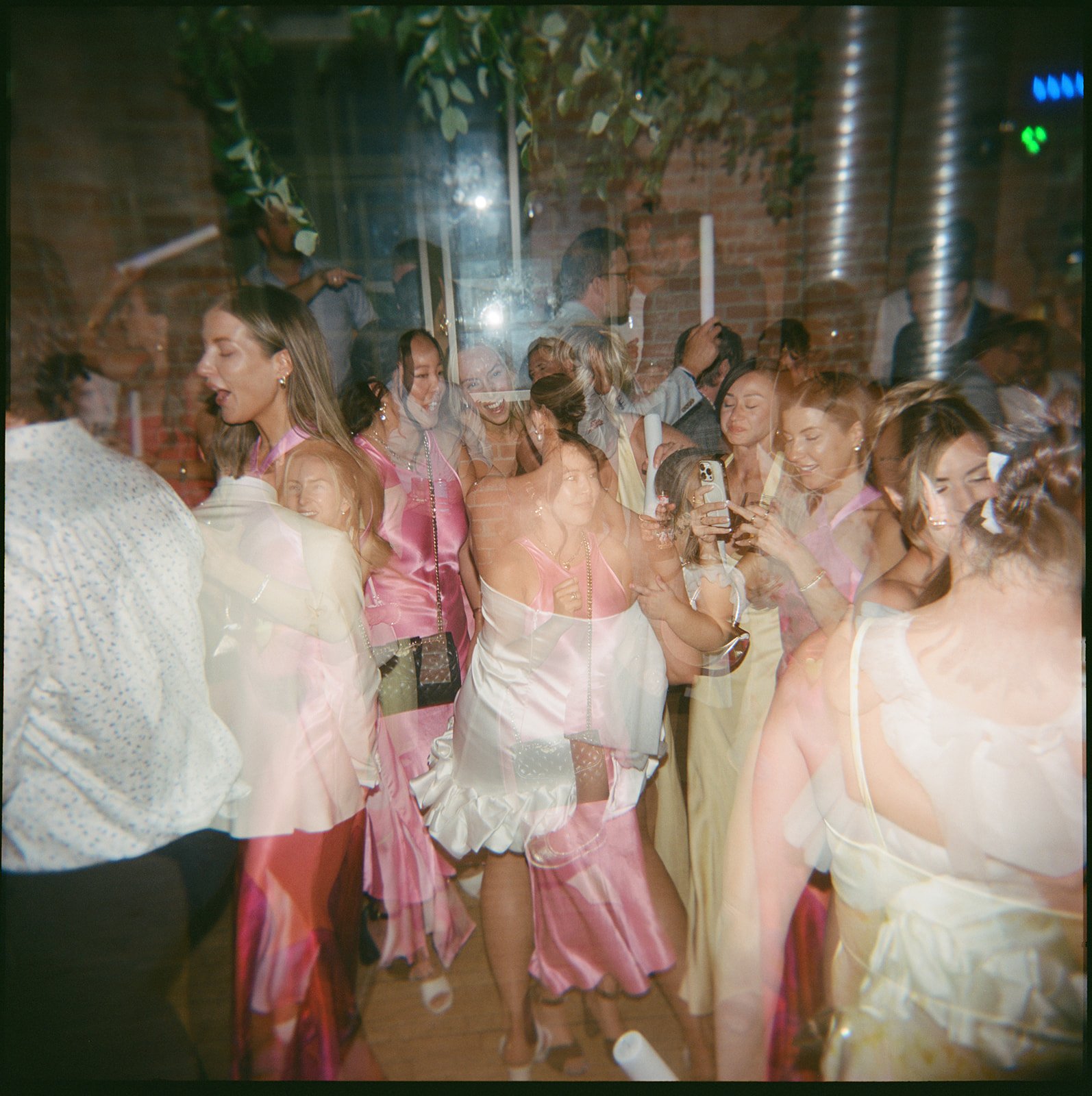 Group of people gathered at a celebration or party, some dressed in pink and yellow dresses, with lively expressions, taking photos and dancing in a decorated indoor space with brick walls.