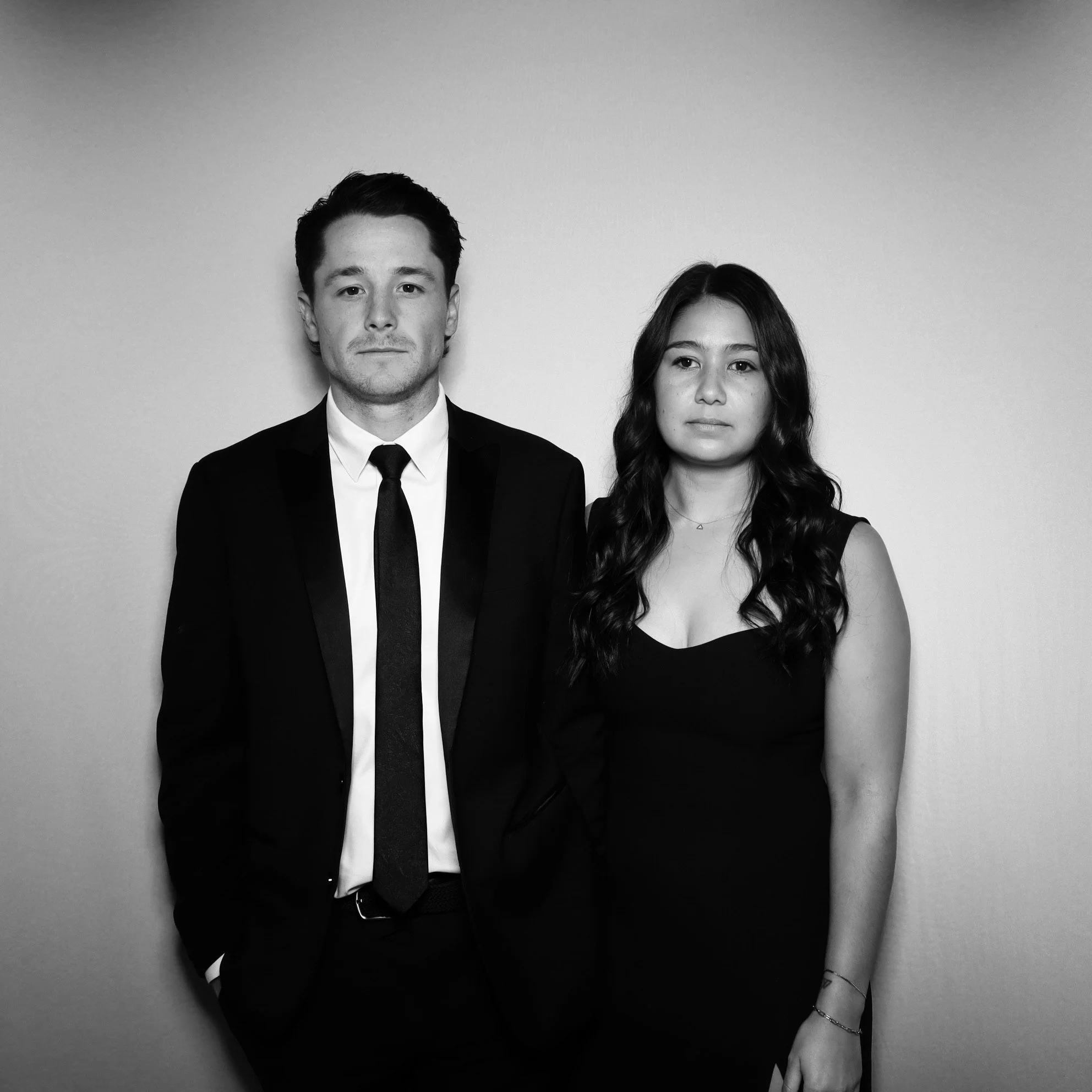 Black and white photo of a man in a suit and a woman in a dress standing against a plain wall, both looking directly at the camera.