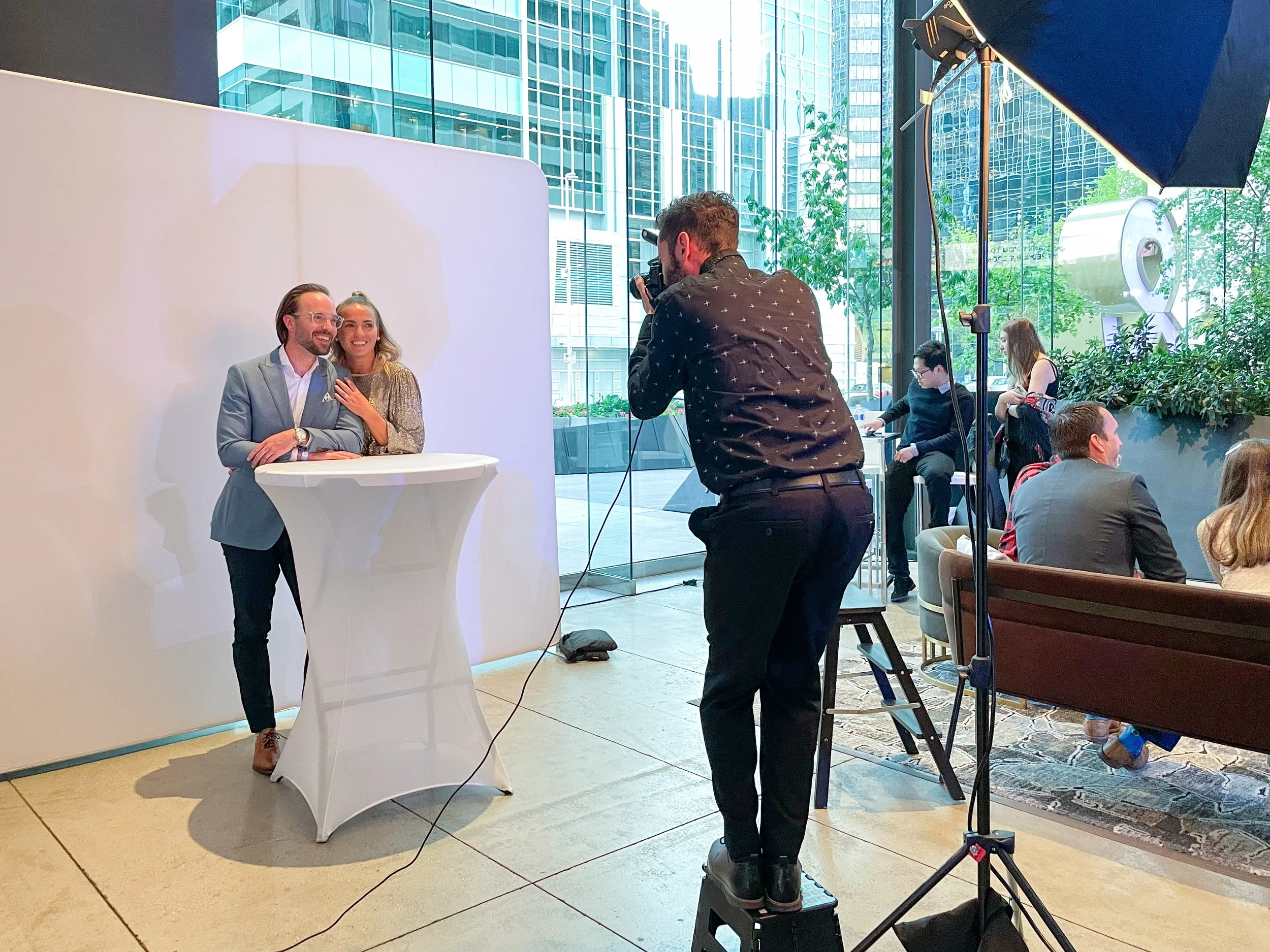 A professional photo shoot with two people at a high-top table in a modern, glass-walled indoor venue. The man and woman are smiling, dressed in formal attire, and posing for the camera. The photographer stands on a step stool, taking their picture. 