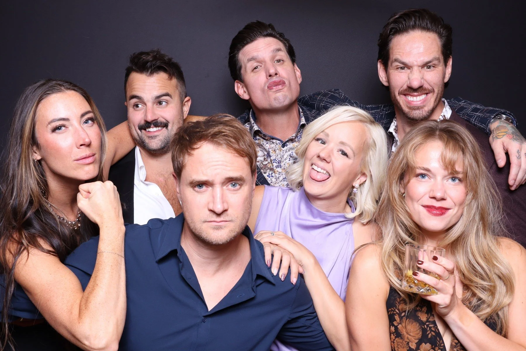 Group of seven adults posing for a photo against a black background, with some making funny faces and others smiling, dressed in party or casual attire.