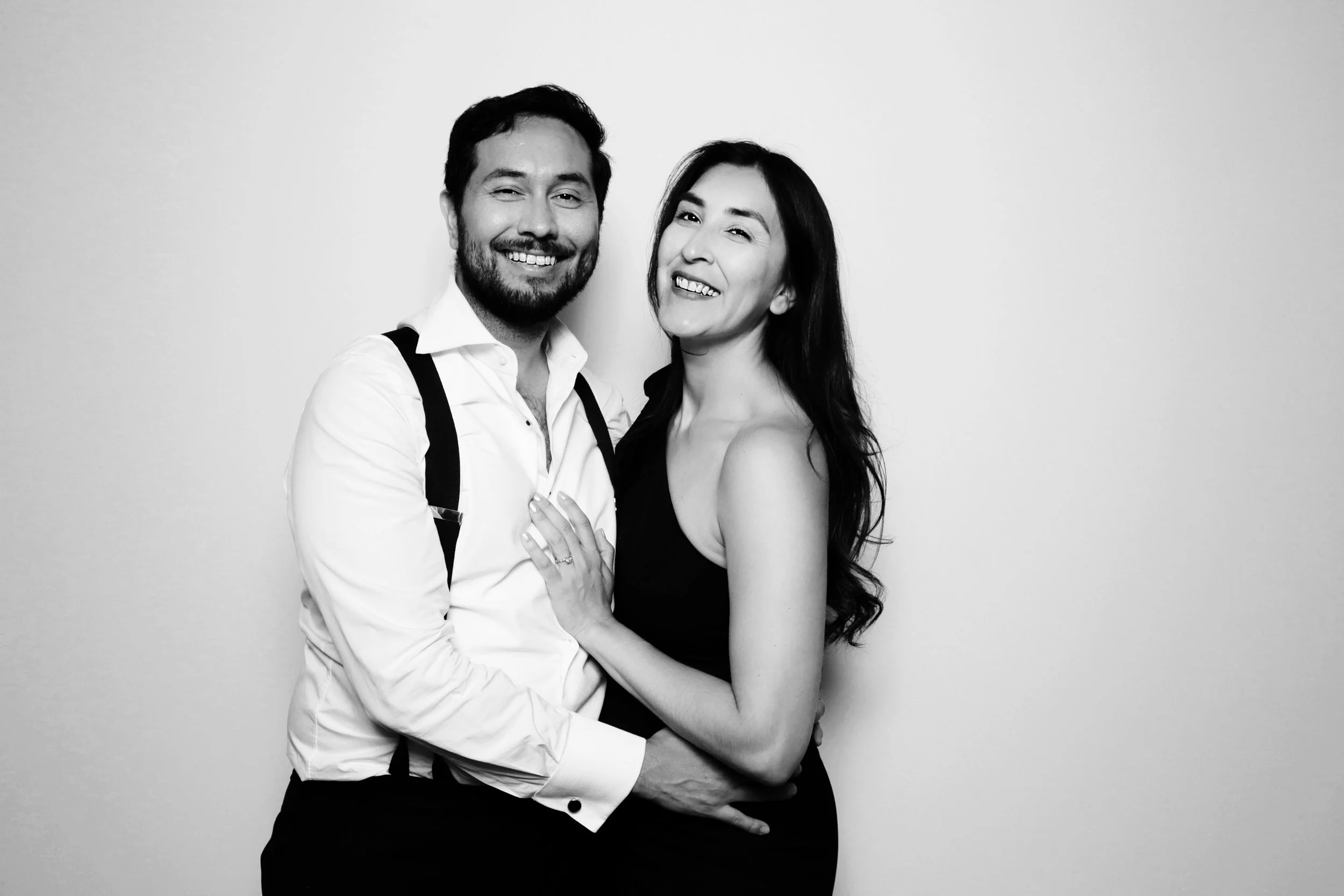 A smiling man and woman standing close together, holding each other, against a plain background. The man has dark hair, a beard, and is wearing a white shirt with suspenders. The woman has long dark hair and is wearing a sleeveless black dress.