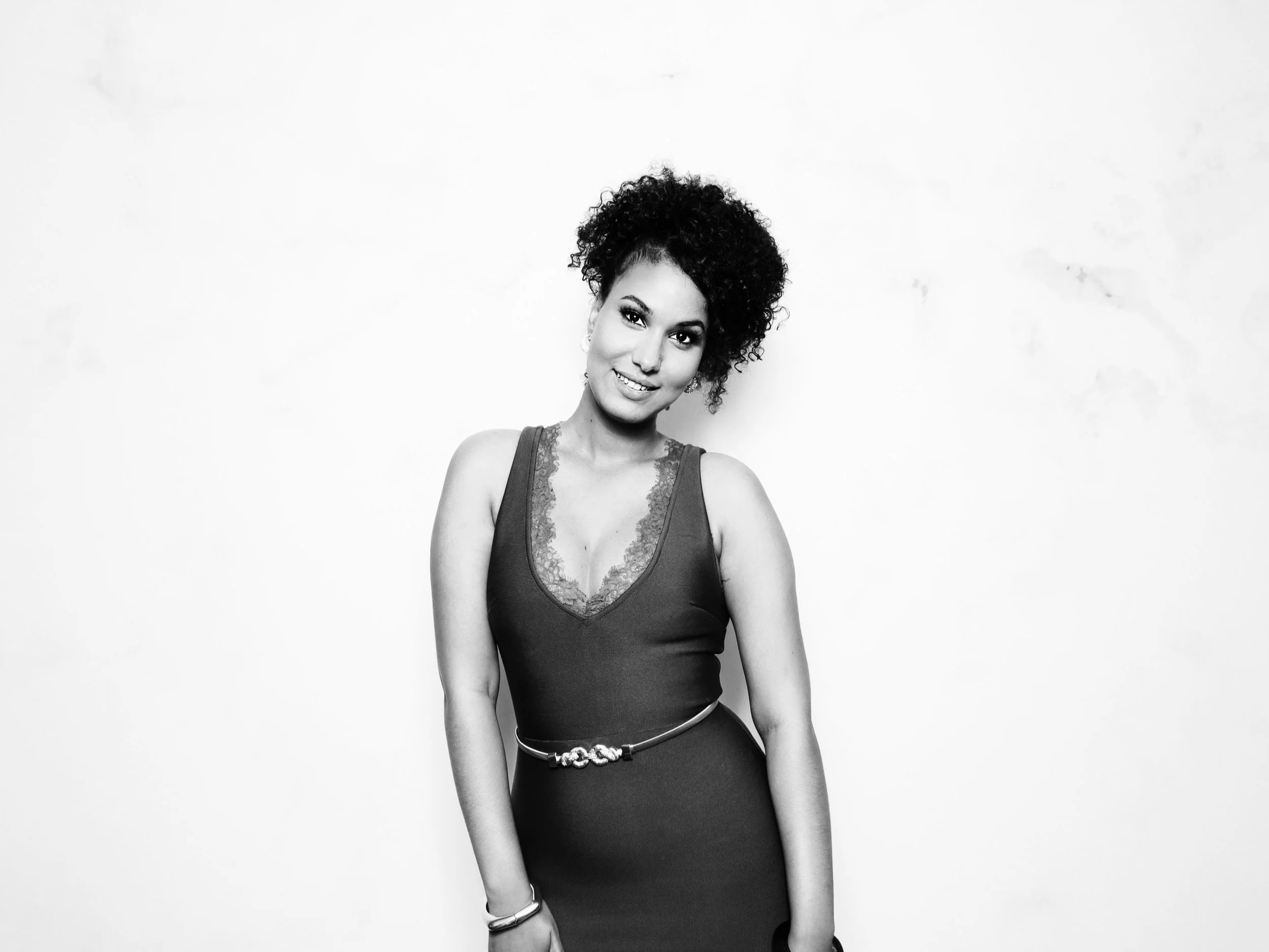 Black and white photo of a smiling woman with curly hair, wearing a sleeveless dress with lace detail and a belt, standing against a plain background.