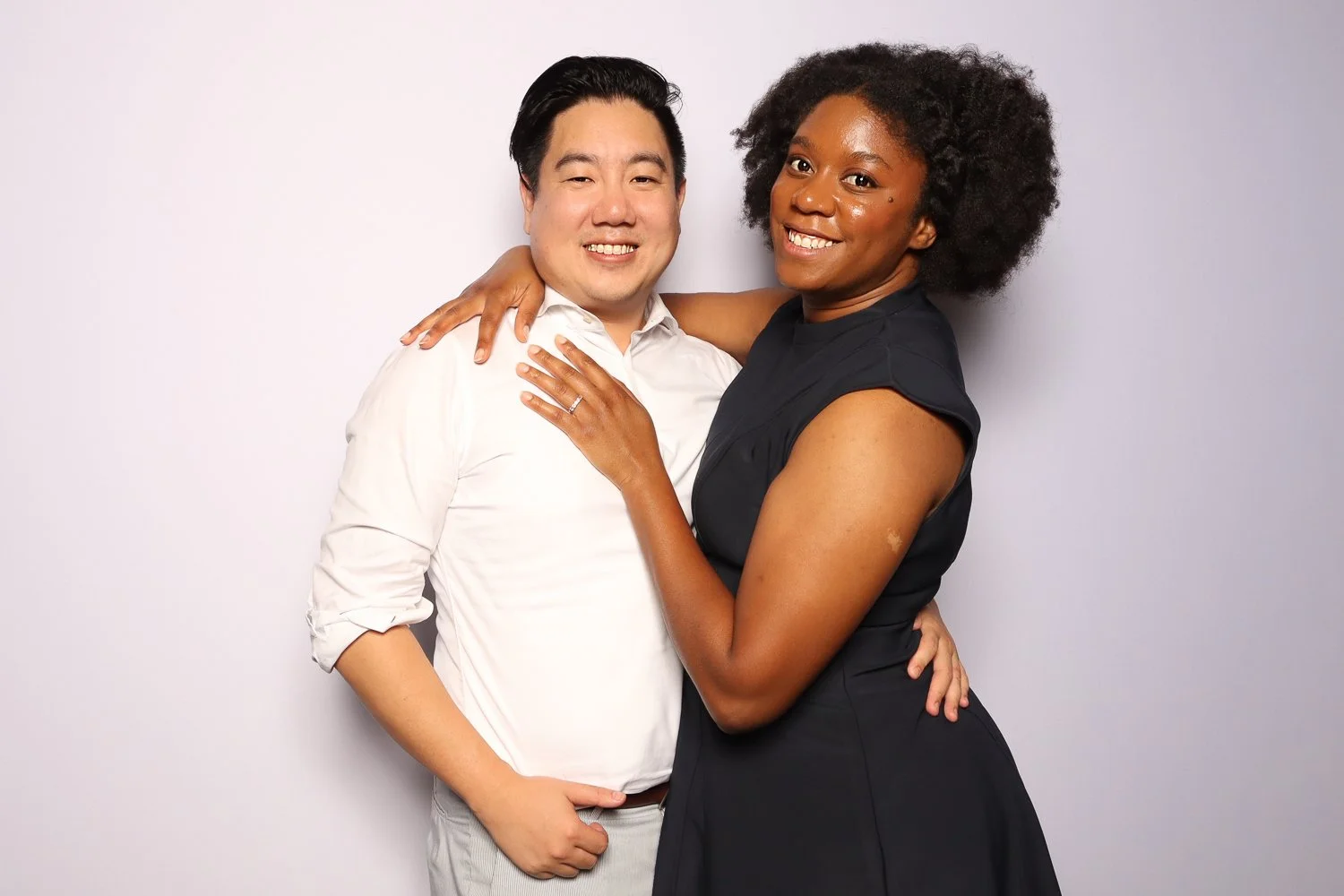 A smiling Black woman with curly hair hugging an Asian man with short black hair against a plain white background.