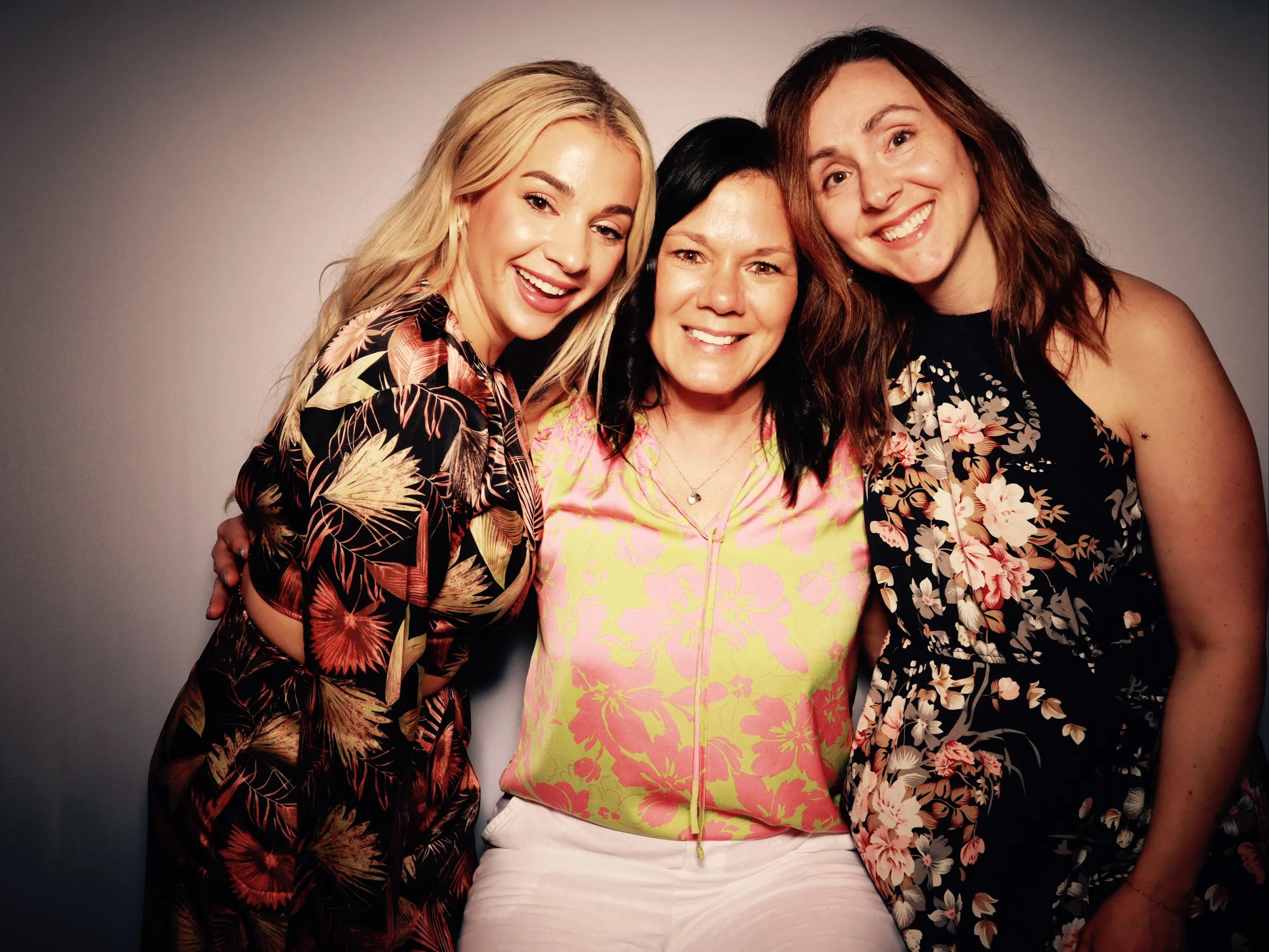 Three women smiling, standing close together, wearing floral-patterned tops, with a plain gradient background.