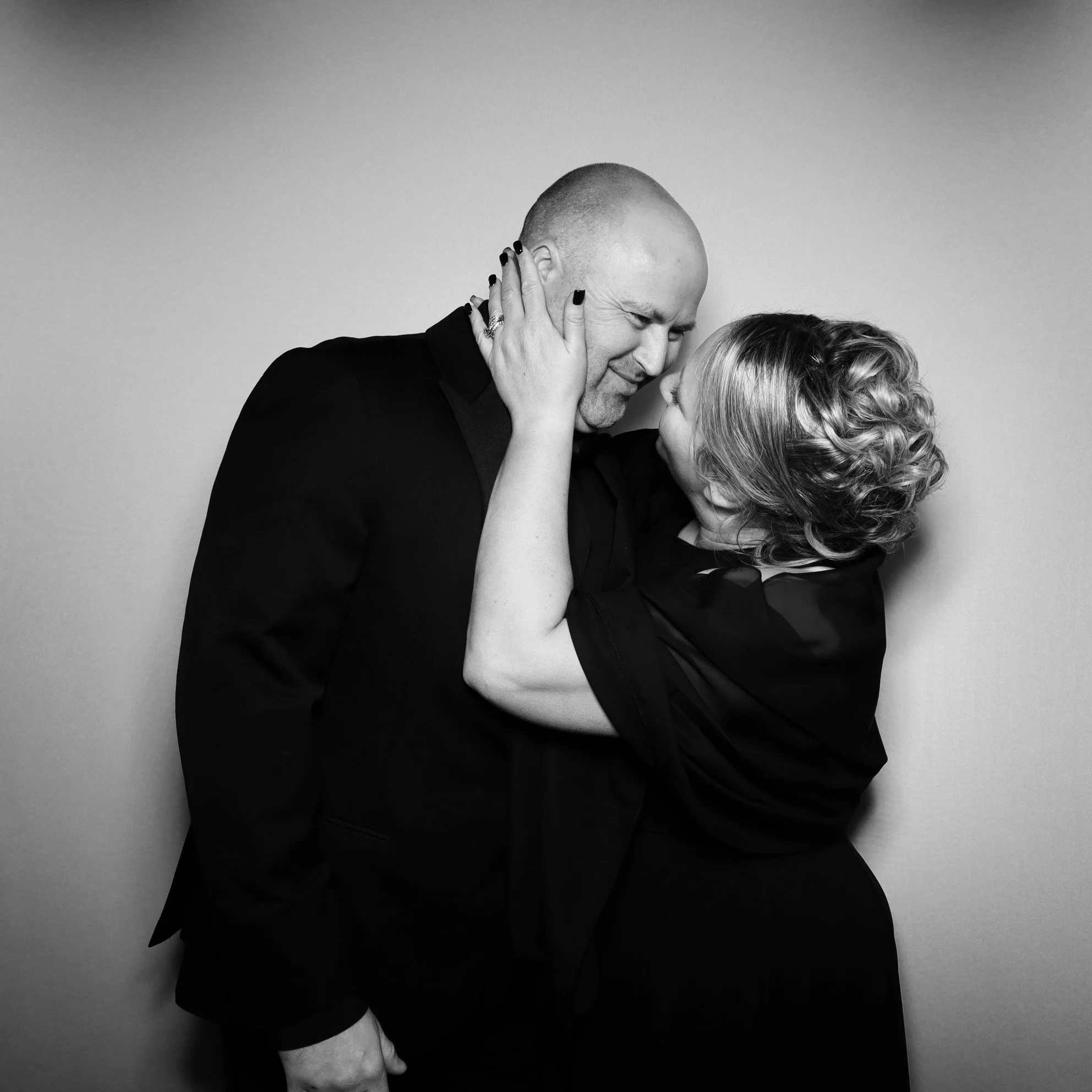 Black and white photo of a man and woman embracing closely, their foreheads touching, and smiling with eyes closed, against a plain background.