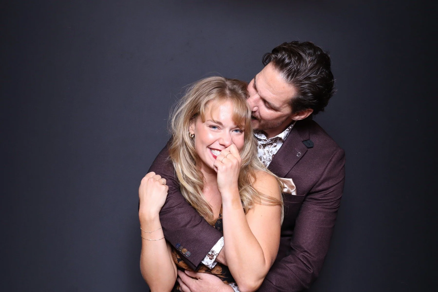 A man and woman sharing a joyful moment, with the man kissing the woman's temple and both smiling; dark background.