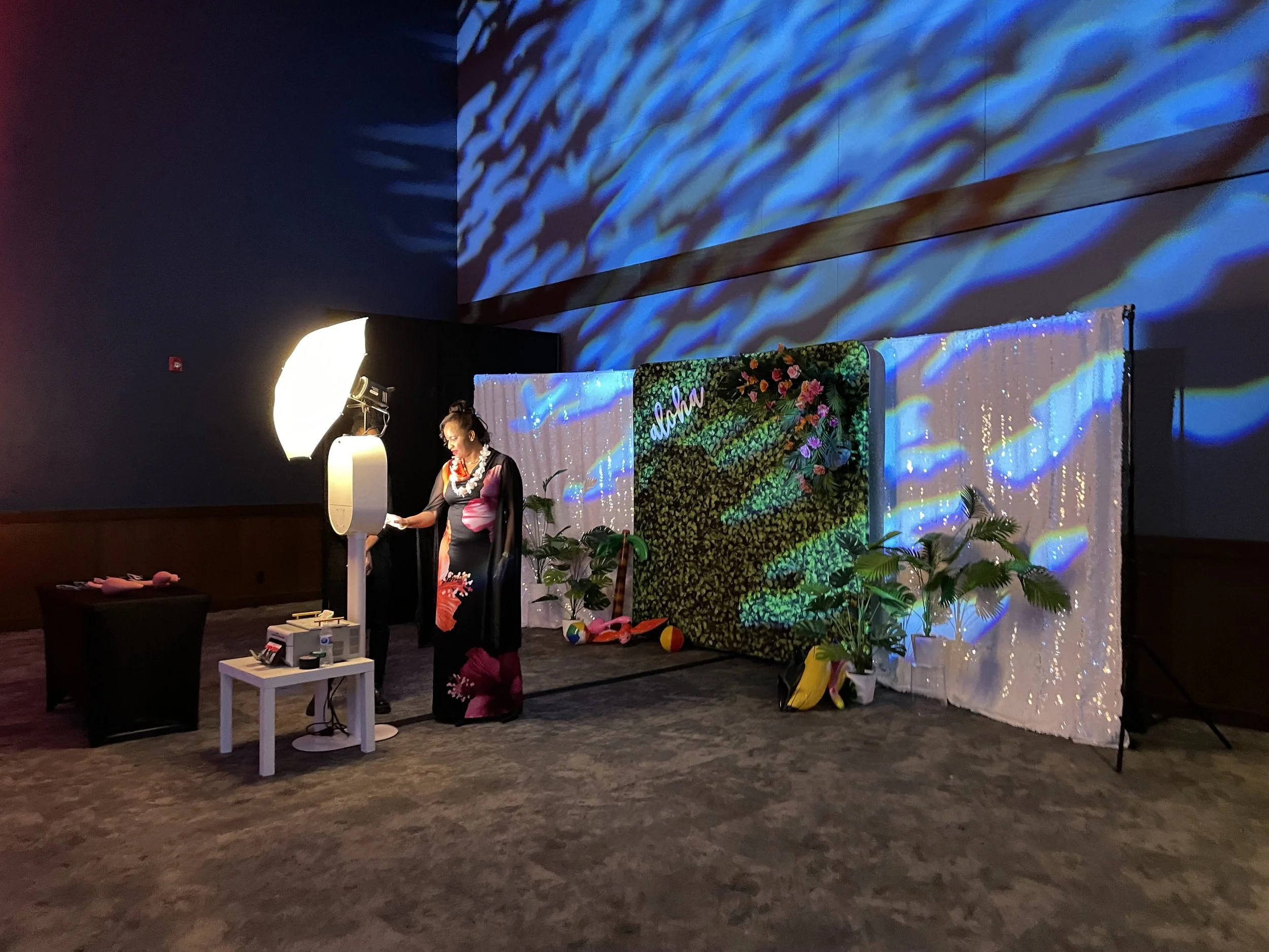 A woman in traditional Hawaiian attire standing in front of a backdrop that says 'Aloha' with plants and beach toys around her, illuminated by studio lighting.