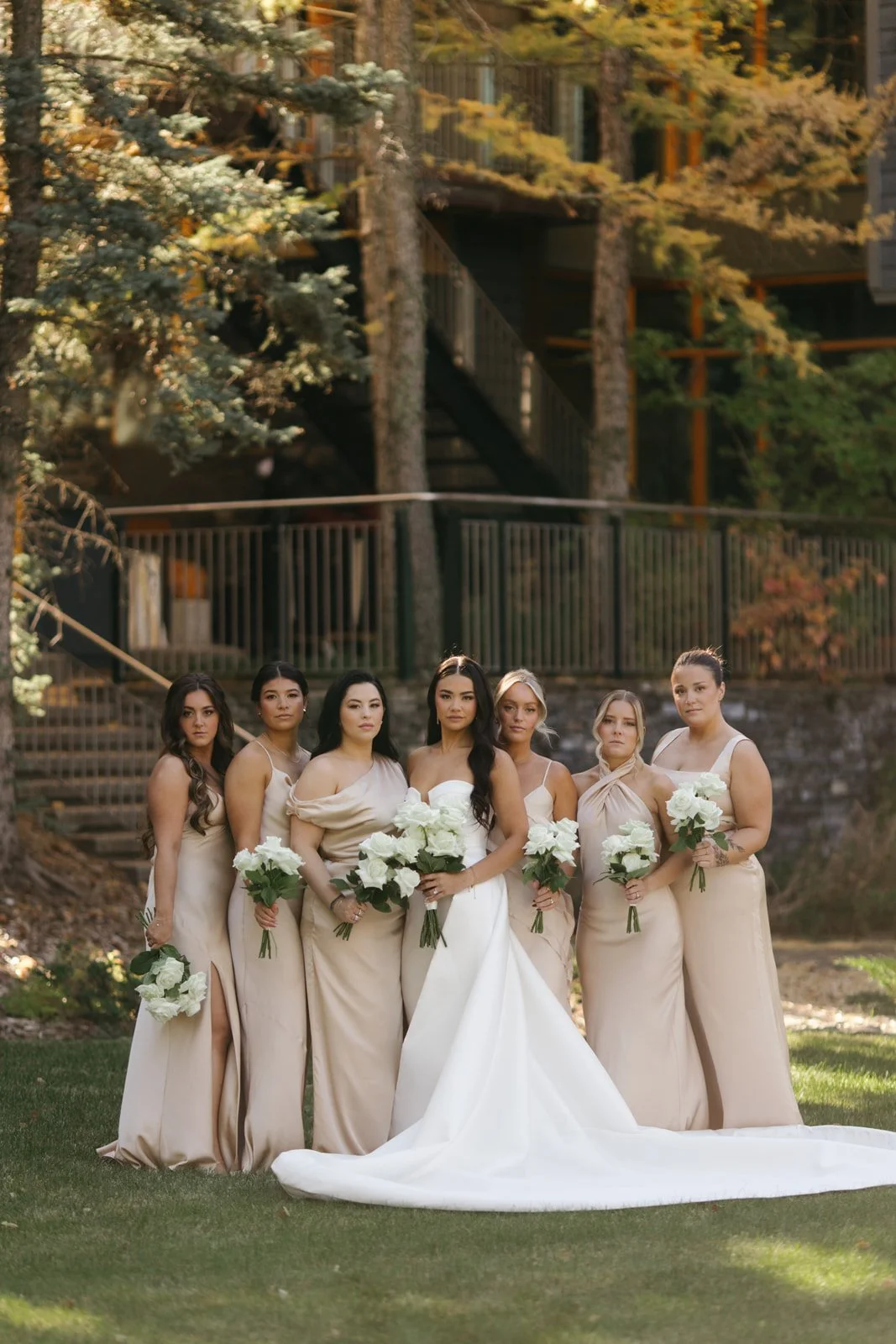 A bride and six bridesmaids standing outdoors on grass, holding white bouquets, with trees and a building with stairs in the background.