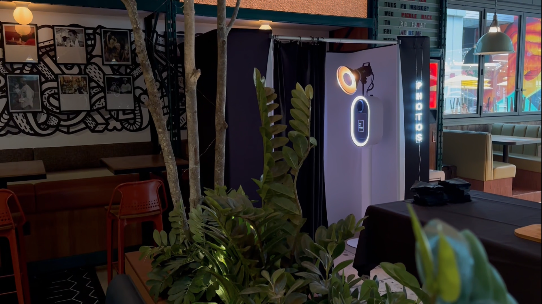 Interior of a restaurant or cafe with a photo display wall, tables, chairs, a photo booth with lighting, and a window showing outside colorful mural artwork.