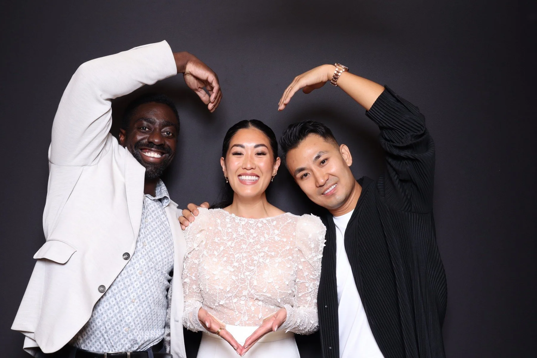 Three diverse friends smiling and making a heart shape with their arms against a black background.