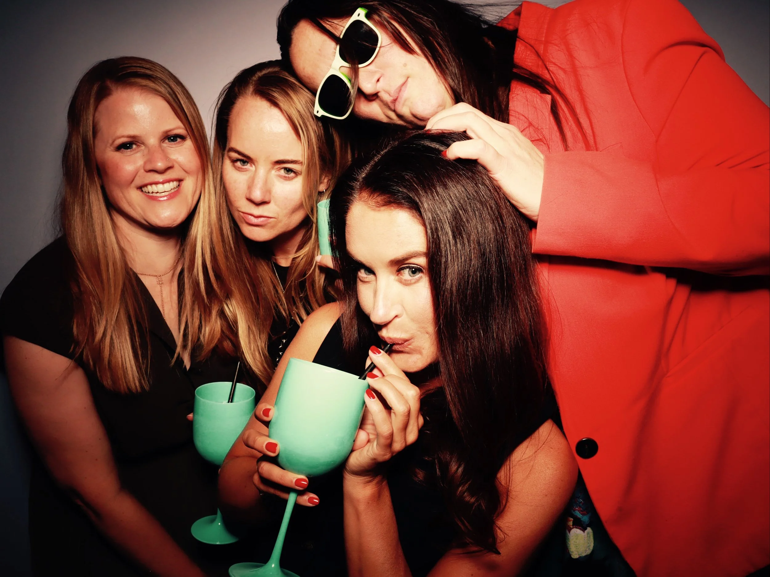A group of five women enjoying a party, holding teal-colored drinks, posing closely together, and smiling. One woman is wearing sunglasses and an orange blazer, while the others have different hairstyles and outfits.