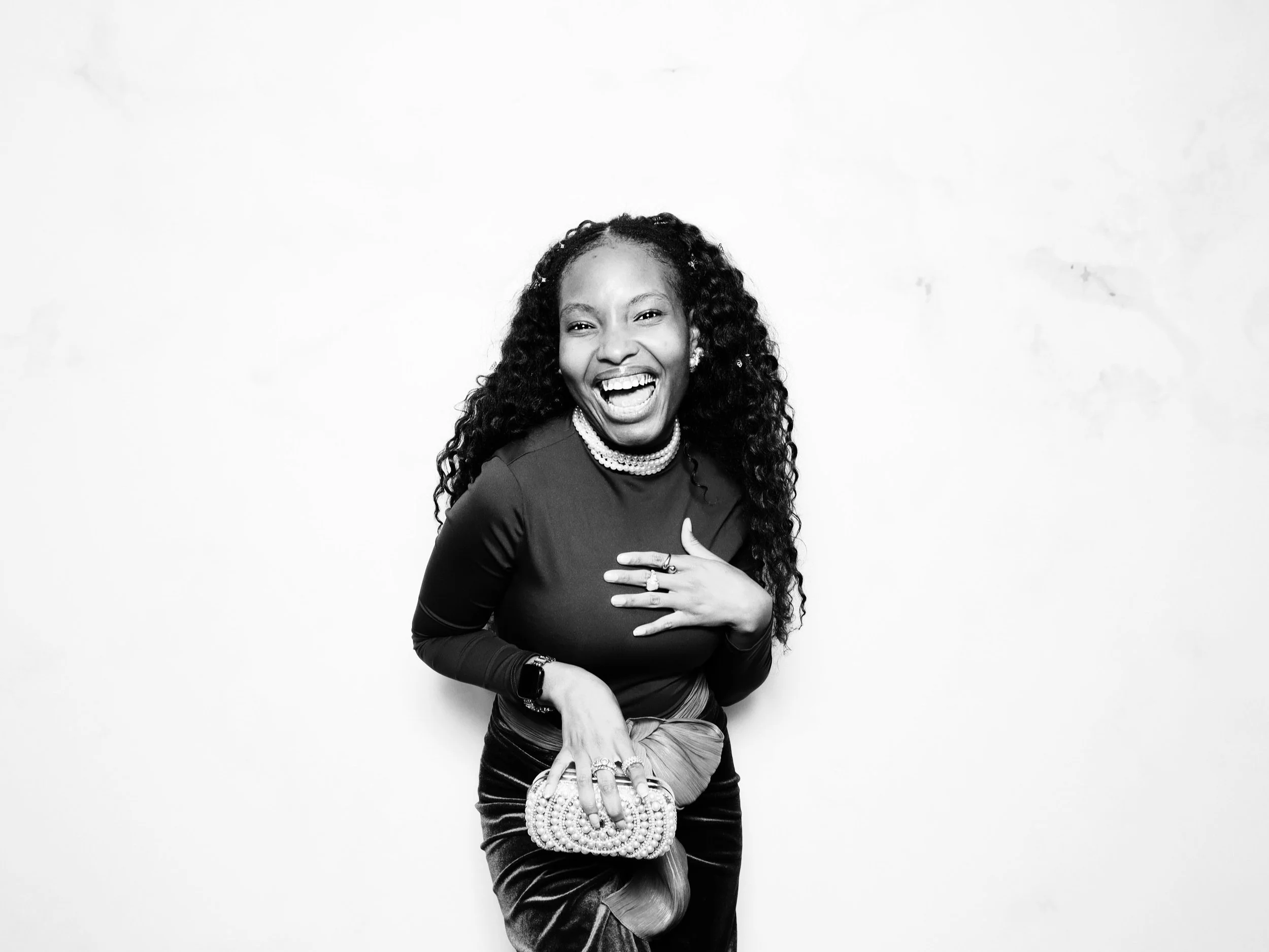 A woman laughing, wearing a long-sleeve top, layered necklaces, holding a beaded clutch, and standing against a plain background.