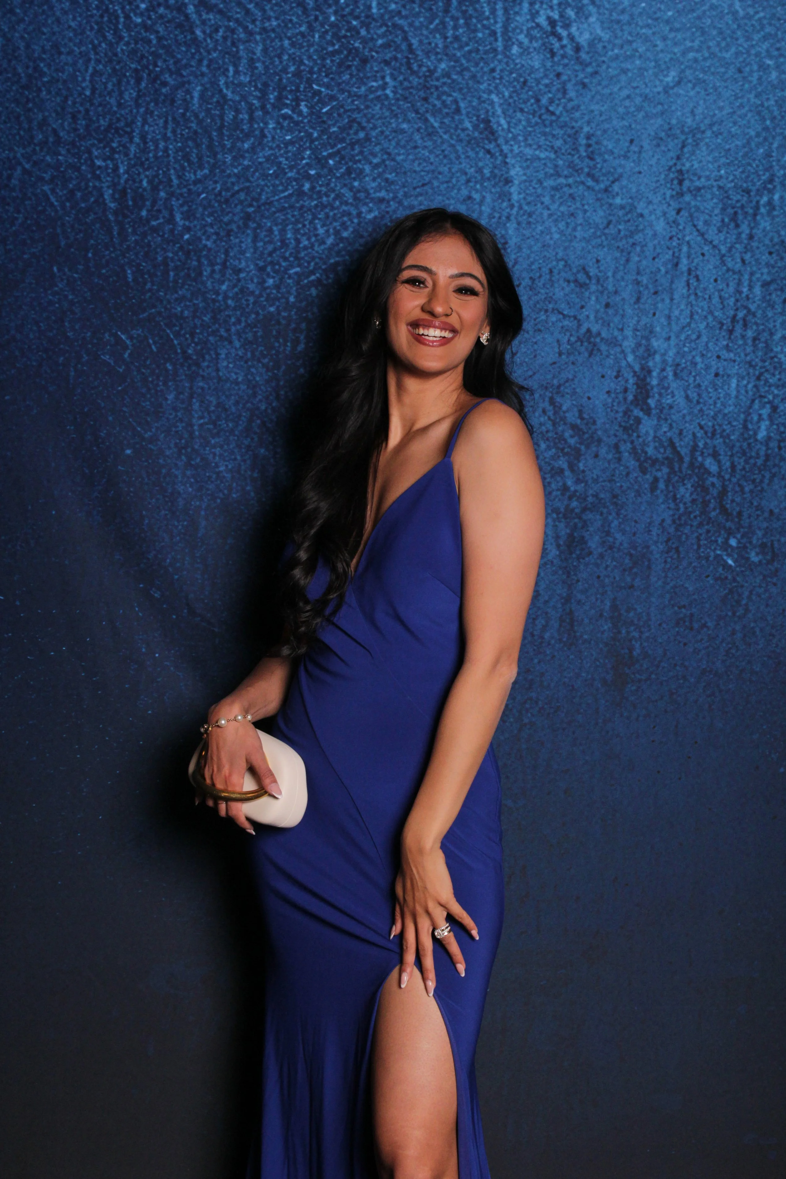 A woman in a royal blue evening gown with a thigh-high slit, smiling against a dark textured blue background, holding a small white clutch.